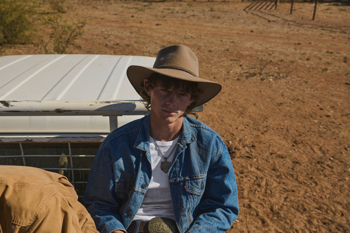 A person wearing a wide-brim brown hat, blue denim jacket, and white t-shirt sitting outdoors in a dry, dusty environment next to a white vehicle.