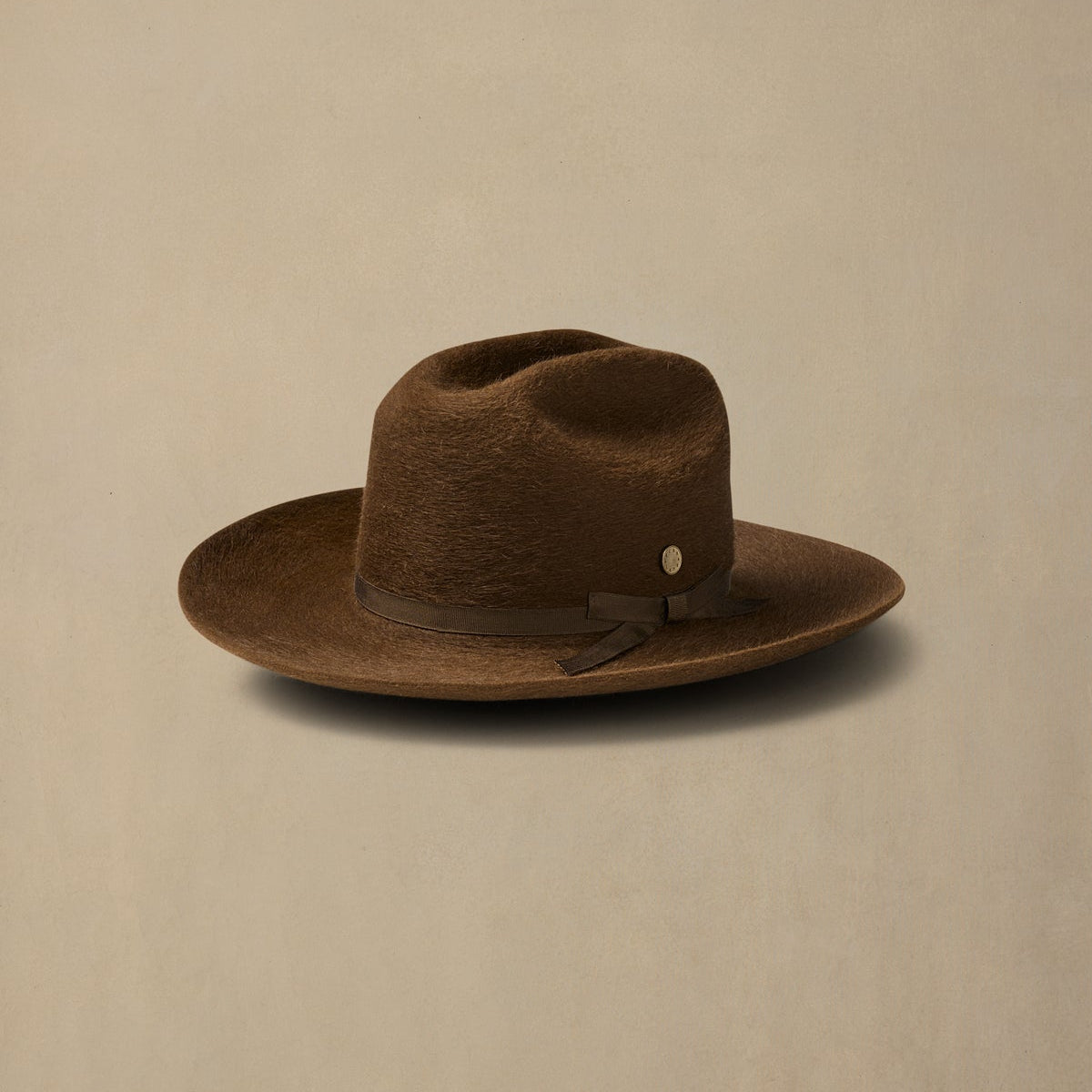 Brown felt cowboy hat with grosgrain band and metal badge on neutral background