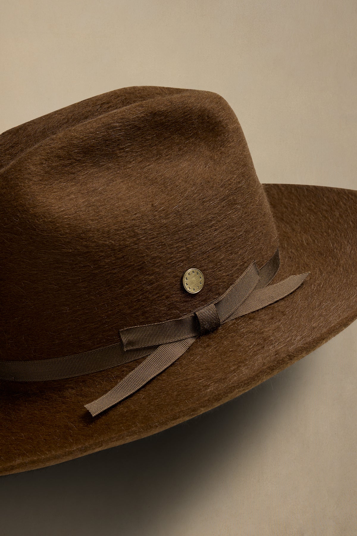 Brown fur felt cowboy hat with grosgrain ribbon band and decorative pin, angled view against neutral background