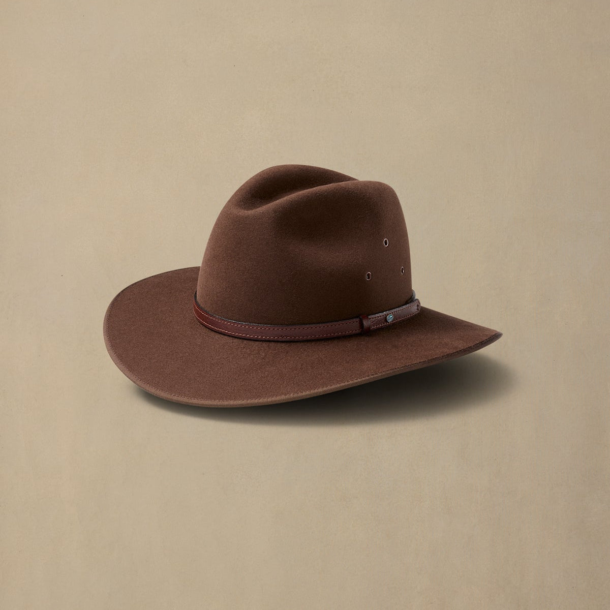 Brown felt cowboy hat with leather band and decorative stitching displayed against neutral beige background