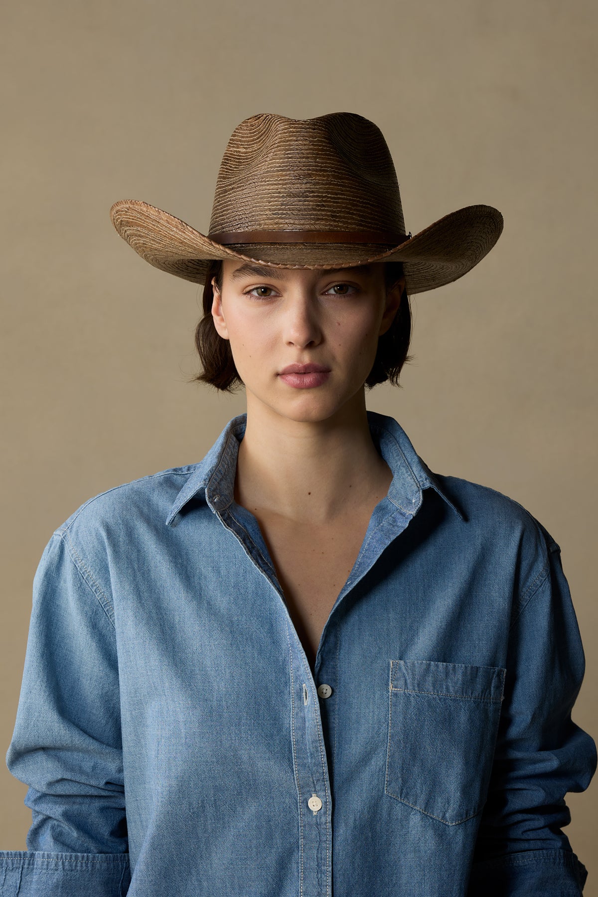 Woman wearing dark toast straw cowboy hat with wide brim and denim shirt against neutral beige background