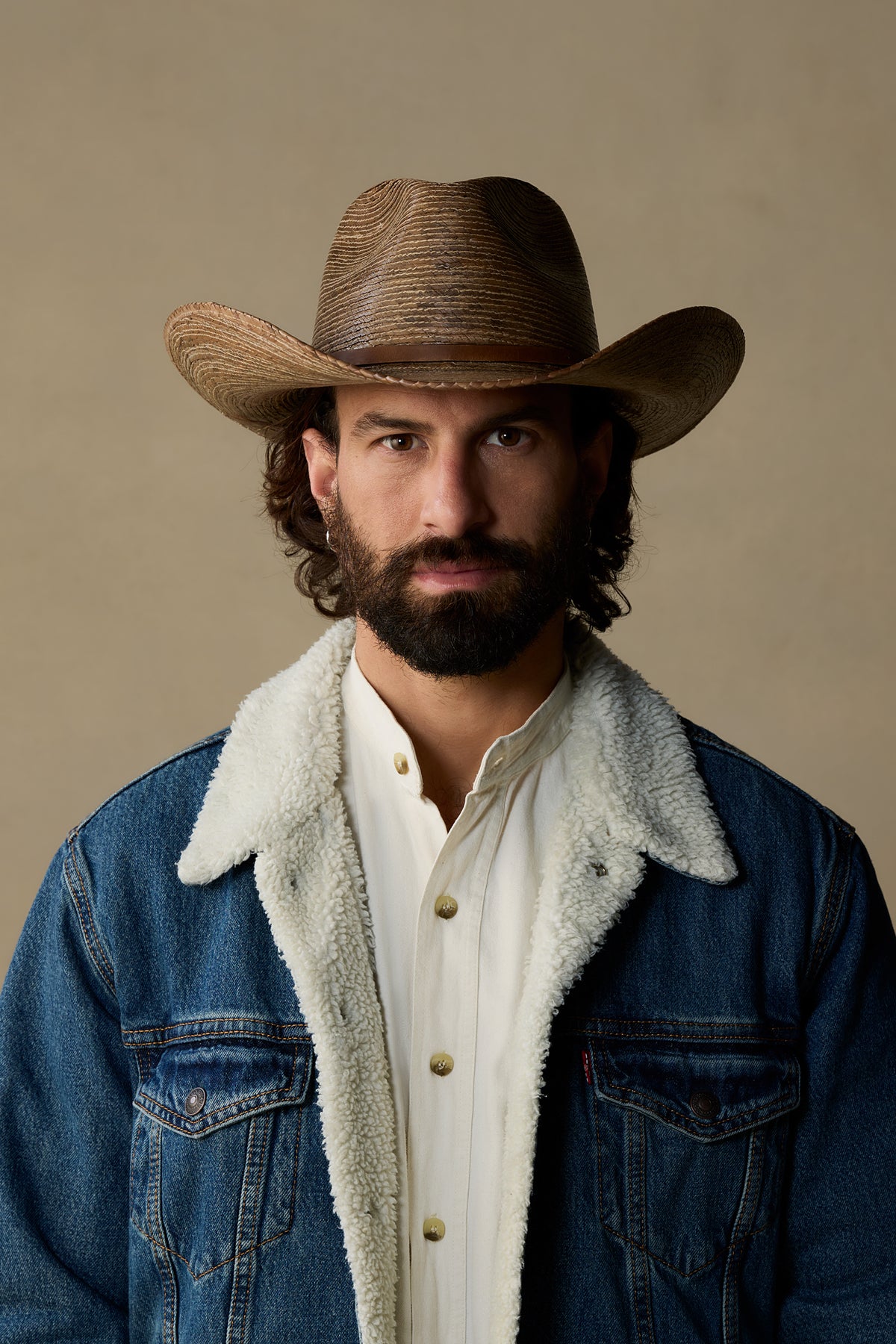 Man wearing tan straw cowboy hat with denim jacket and sherpa collar against neutral background