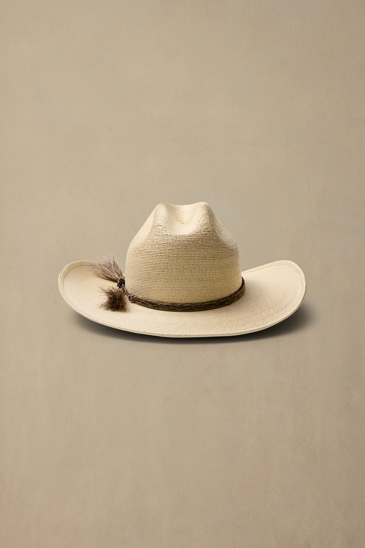 A light beige woven cowboy hat with a dark band and tasselaccent, photographed against a solid neutral beige background.