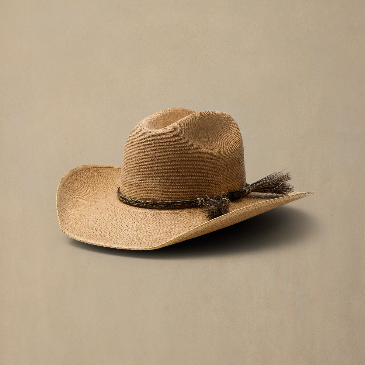 A dark tan woven cowboy hat with a dark brown decorative band and tassel accent, photographed on a solid beige background.
