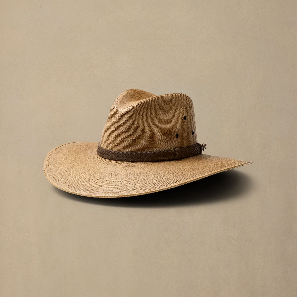 A dark tan woven cowboy hat with a dark brown braided band, photographed on a solid beige background.