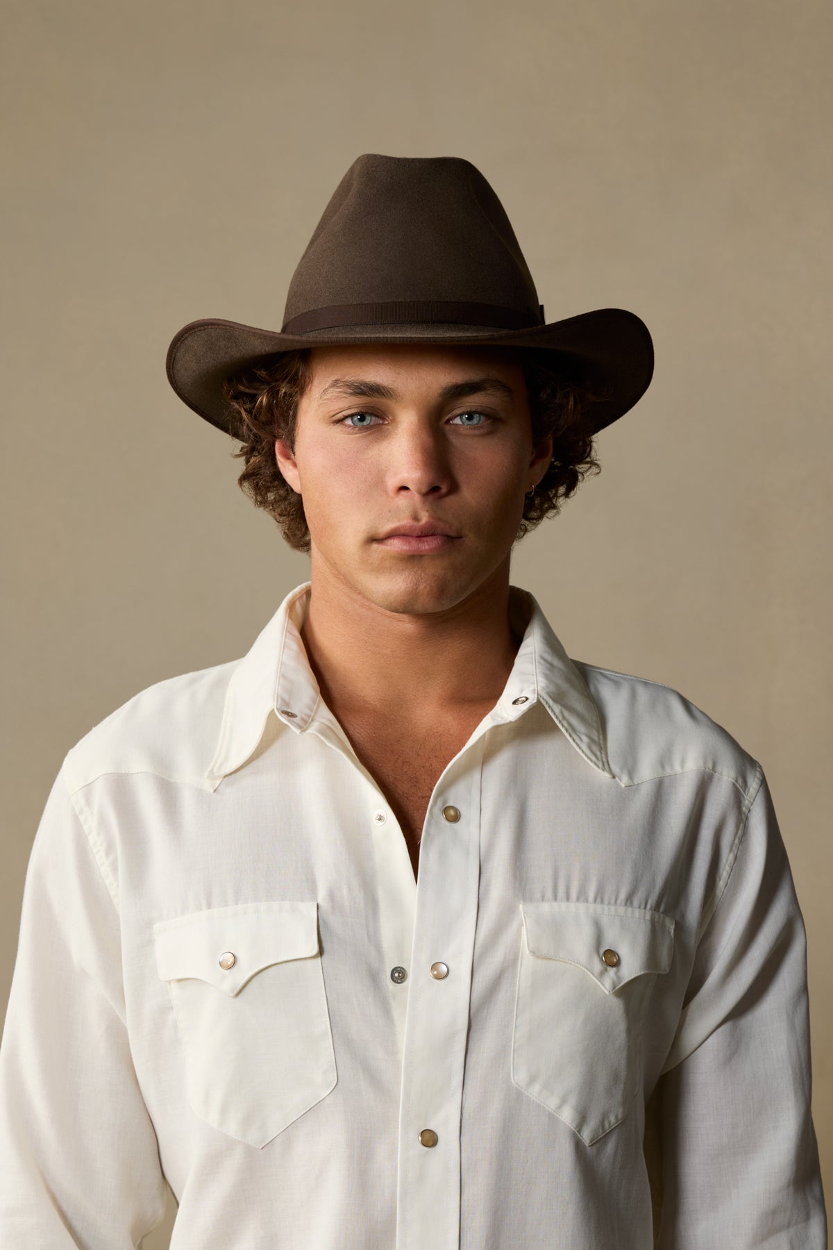Man wearing mid brown cowboy hat with cream western shirt against neutral background