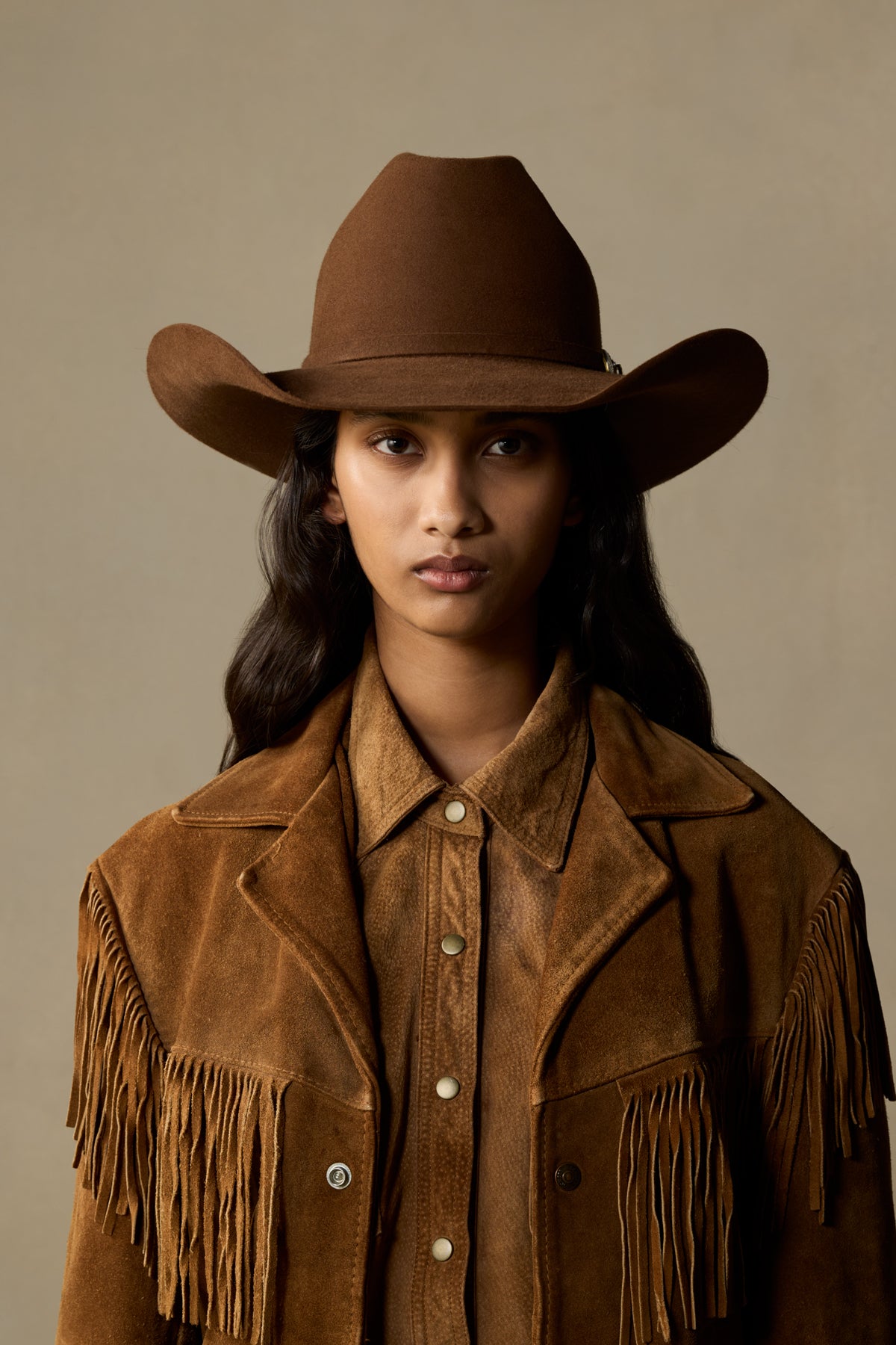 Brown fur felt cowboy hat with wide brim and fringe detailing, worn by model with long dark hair against neutral background