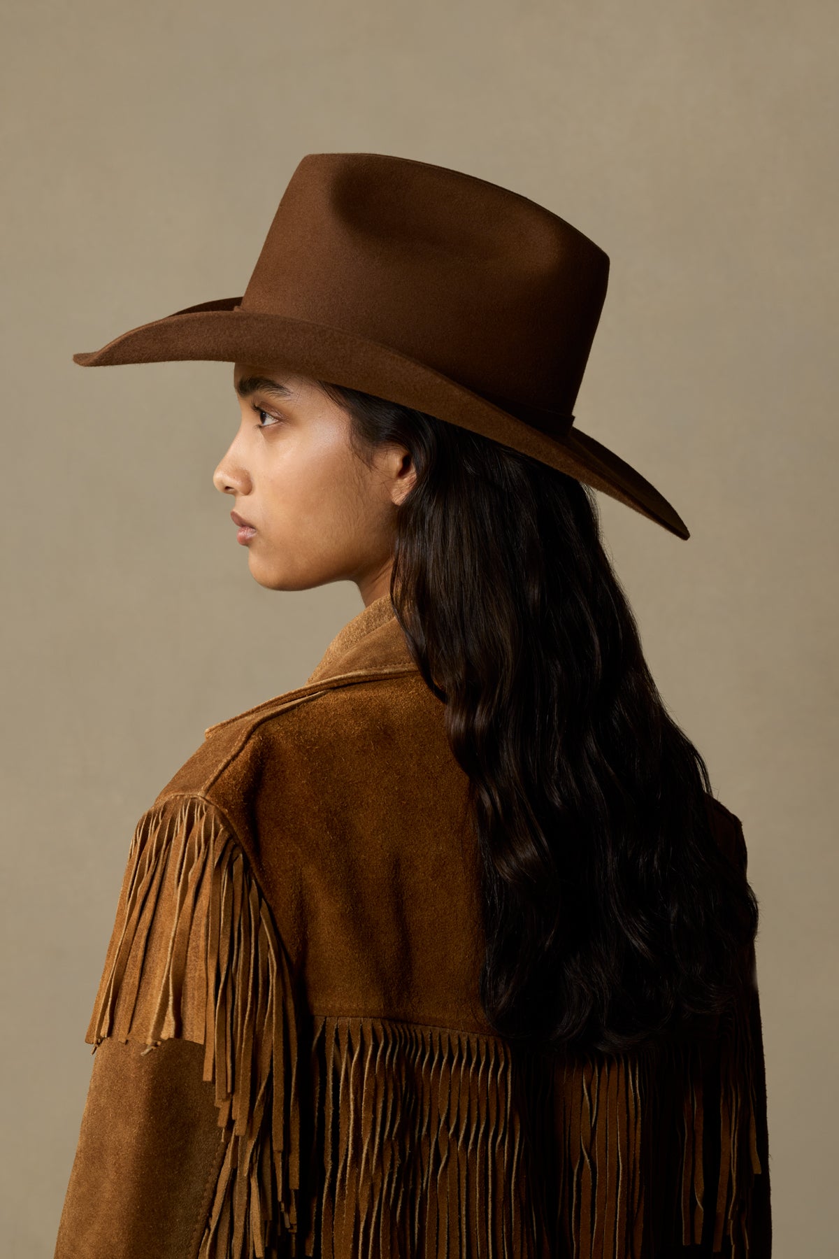 Woman wearing brown fur felt Big Sky cowboy hat with fringe suede jacket, profile view against neutral background