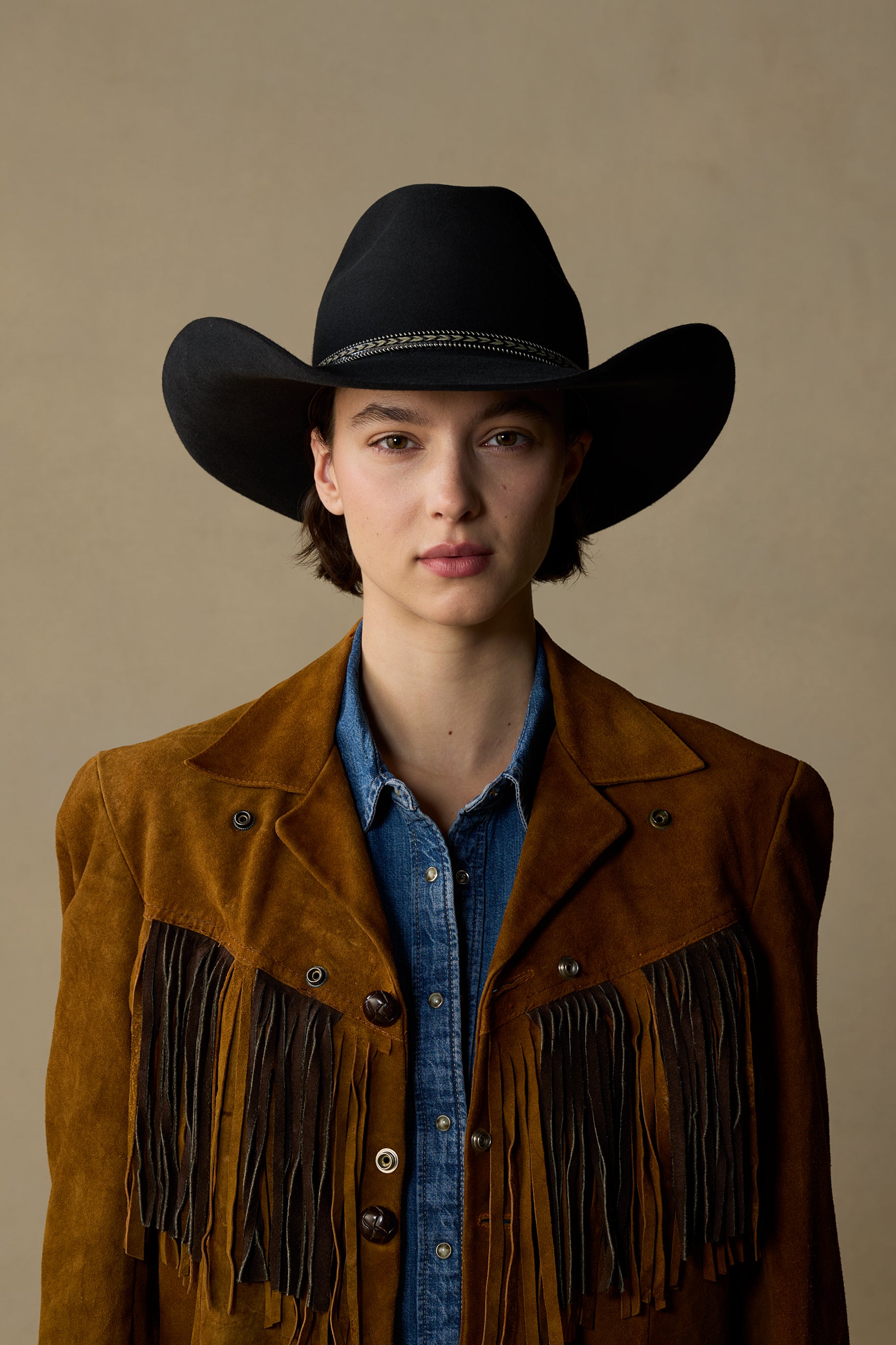 Black cowboy hat with decorative band on model wearing brown fringed suede jacket and denim shirt against neutral background