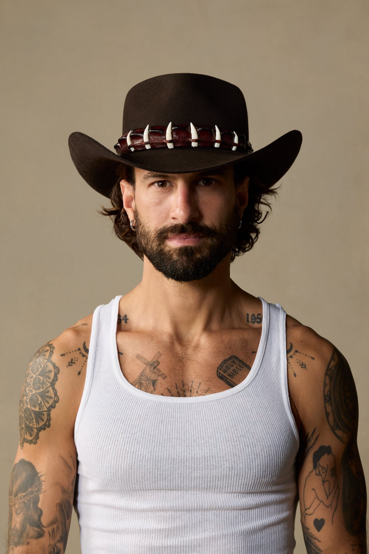 Black cowboy hat with decorative studded band worn by man with full beard and multiple tattoos against neutral background