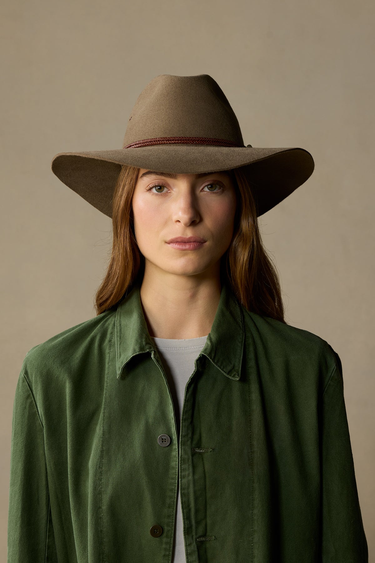 Woman wearing tan fur felt cowboy hat with burgundy band and green jacket against neutral background