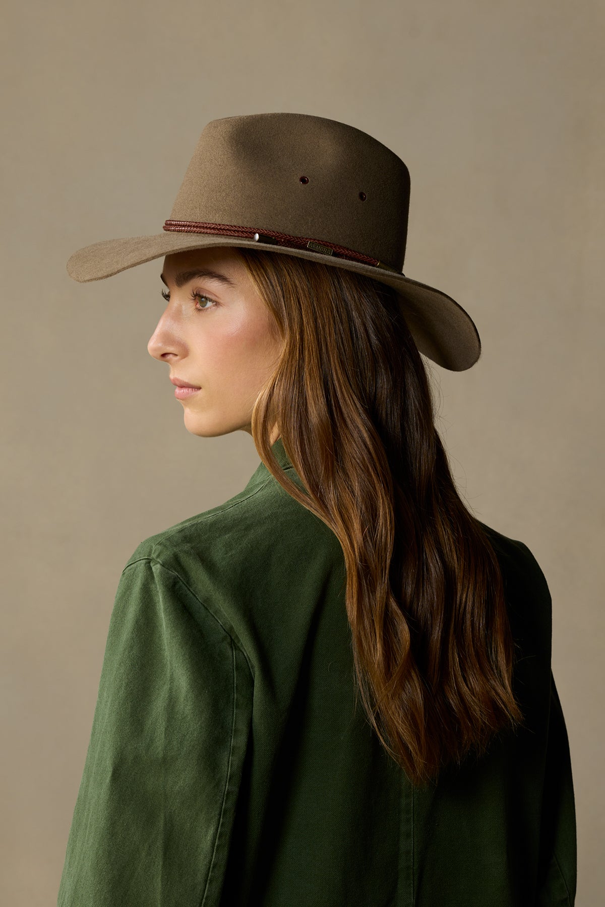Woman wearing tan fur felt cowboy hat with burgundy band and green jacket, side profile view against neutral background