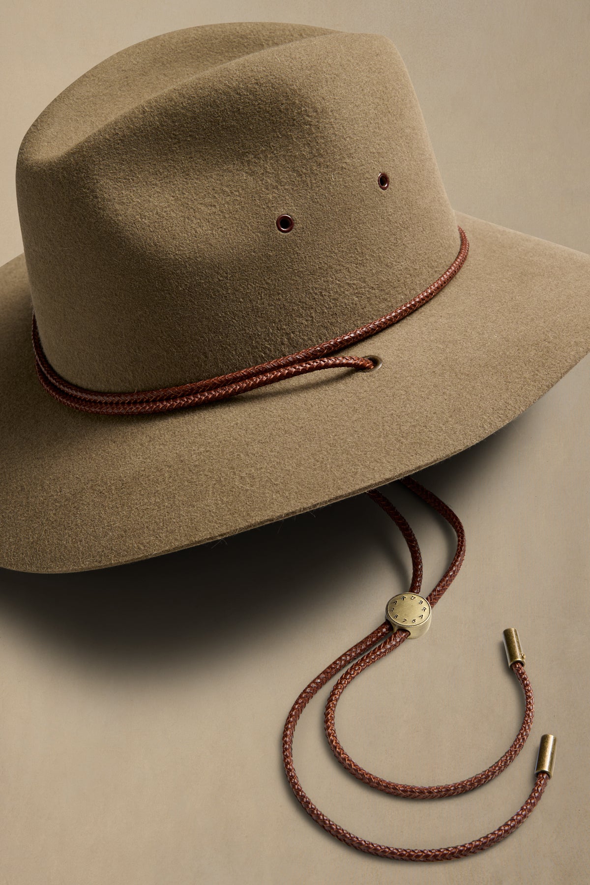 Tan rabbit fur traveller hat with burgundy cord chin strap and metal slide adjuster on cream background