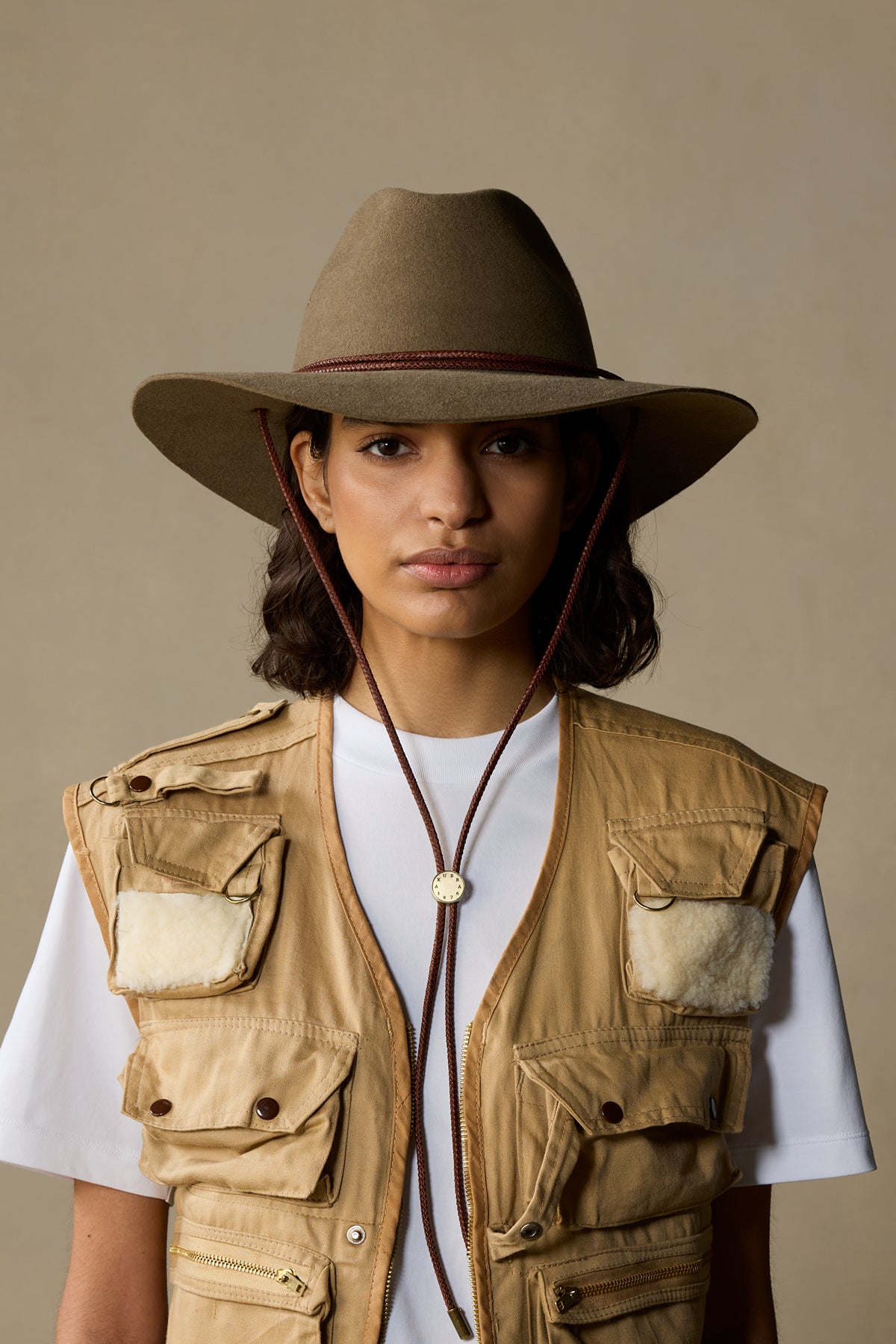 Woman wearing tan rabbit fur traveller hat with chin strap and matching khaki vest over white shirt