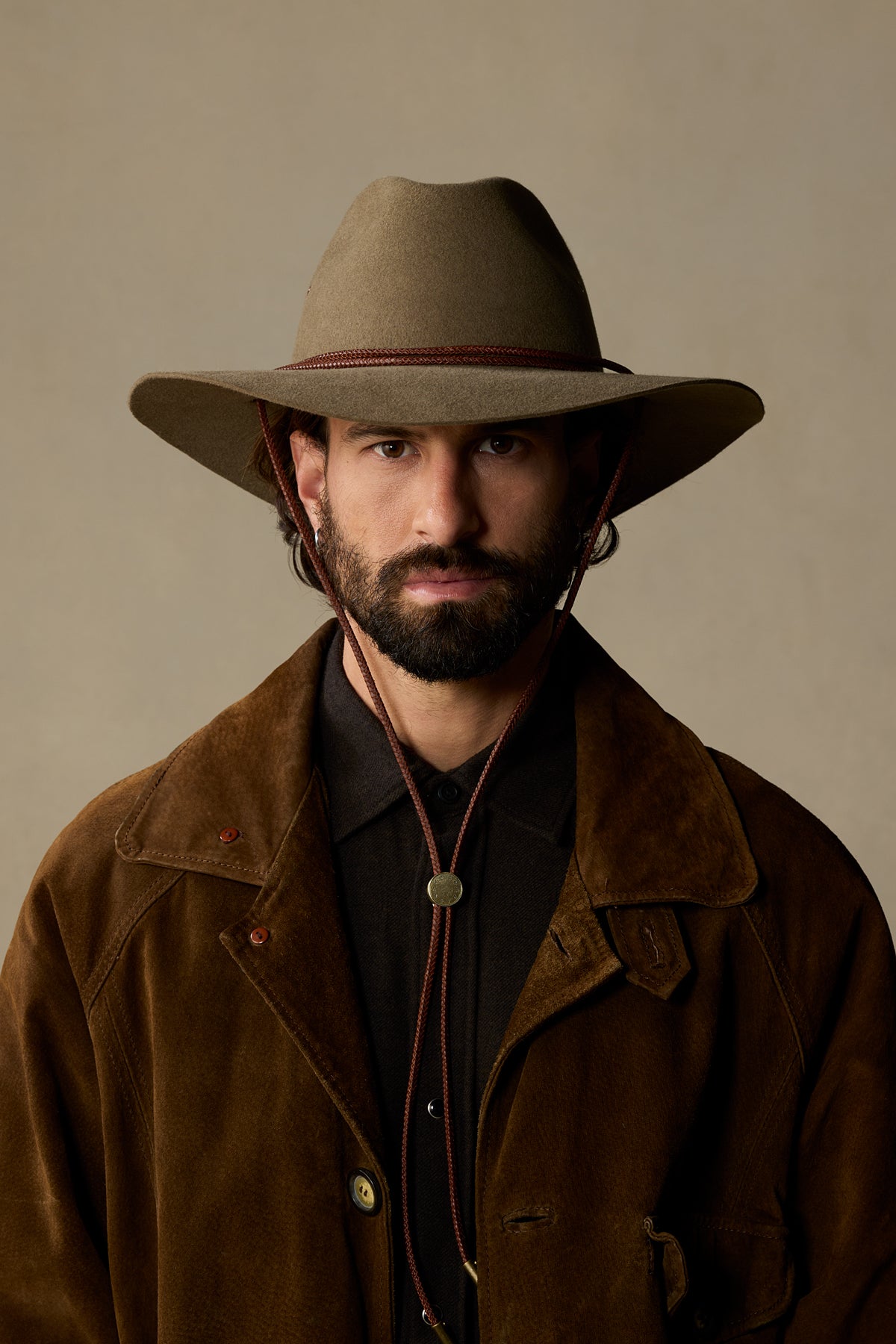 Tan rabbit fur cowboy hat with braided leather chin strap worn by bearded man in brown suede jacket