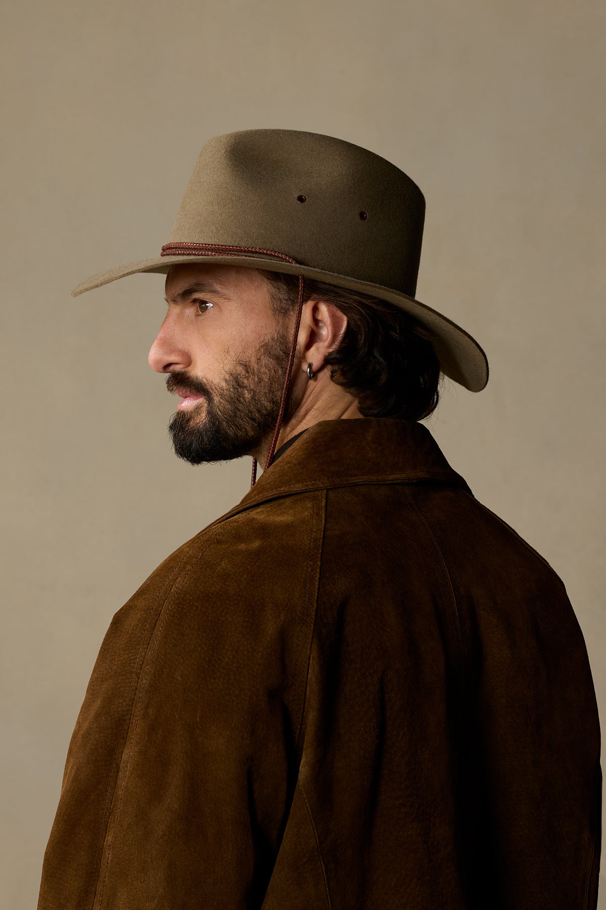 Bearded man wearing tan felt cowboy hat and brown suede jacket, shown in profile against neutral background