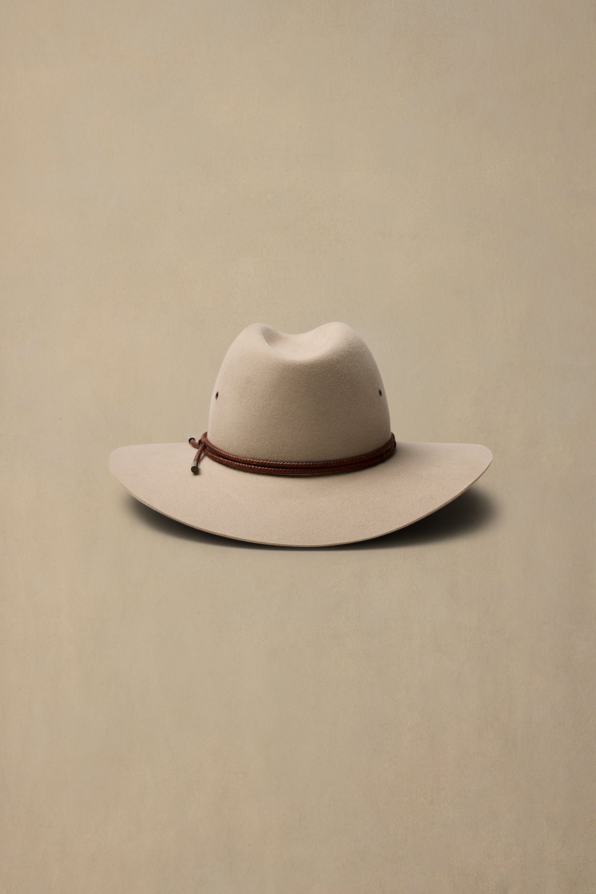 Cream-colored rabbit fur felt cowboy hat with brown cord band on neutral beige background