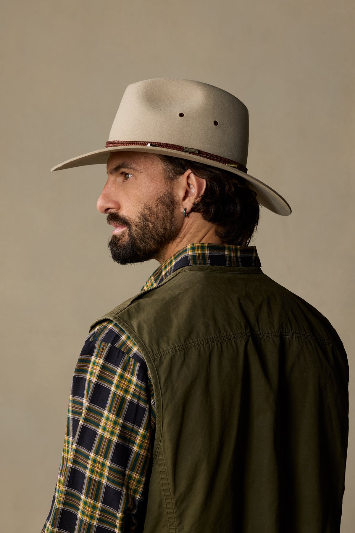 Man wearing sand-colored Traveller cowboy hat with plaid shirt and olive jacket, shown in profile view