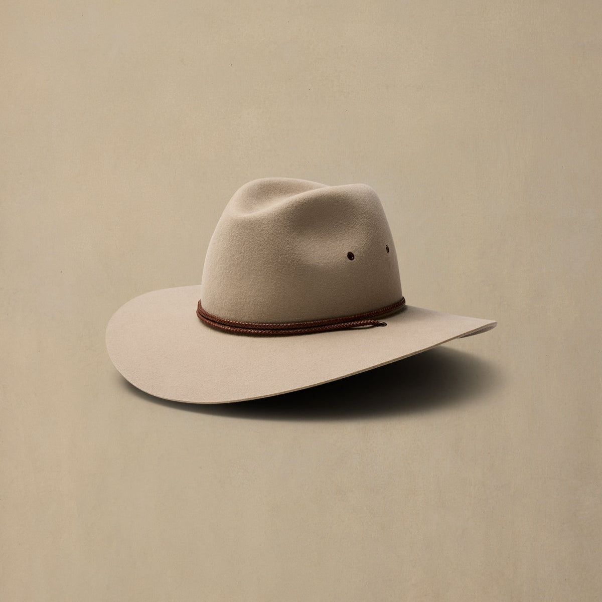 Cream-colored felt cowboy hat with brown braided band, displayed against neutral beige background