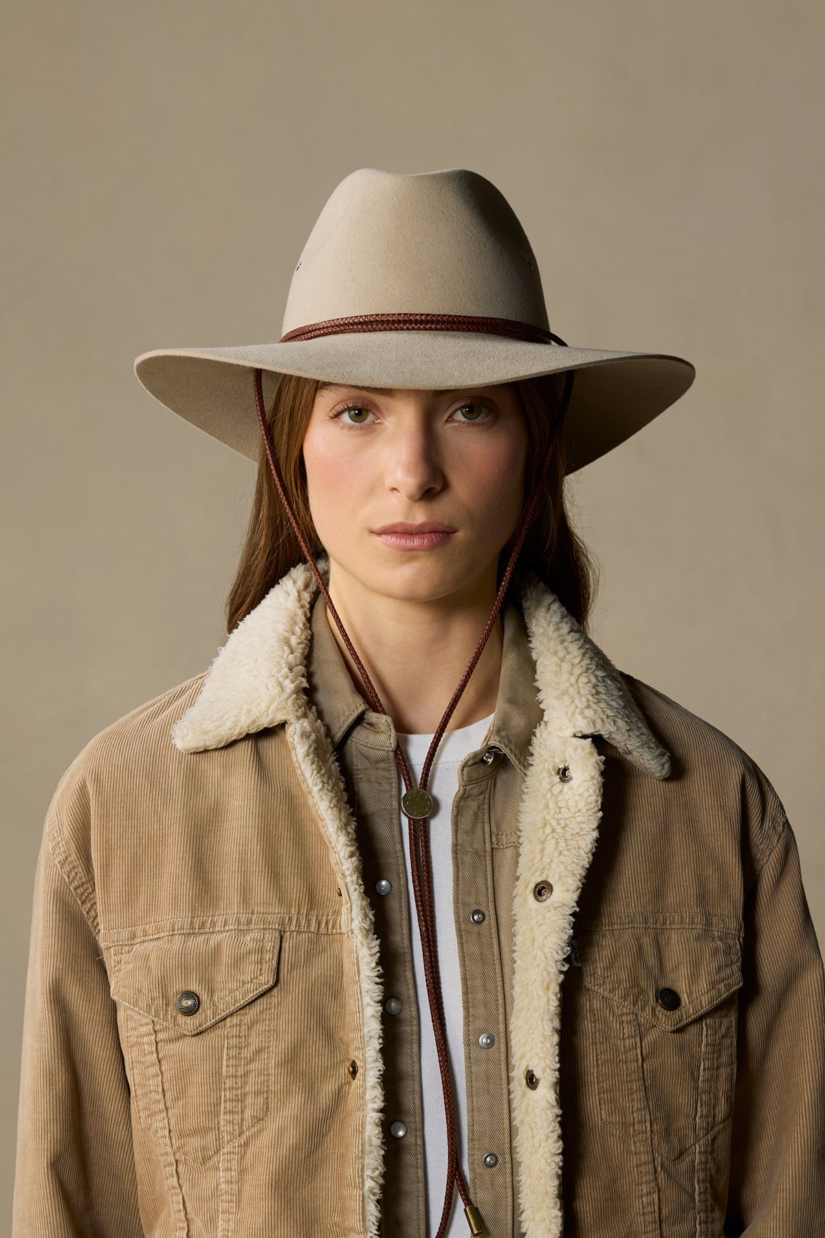 Woman wearing cream felt Traveller hat with burgundy chin cord and sherpa-lined tan corduroy jacket against neutral background