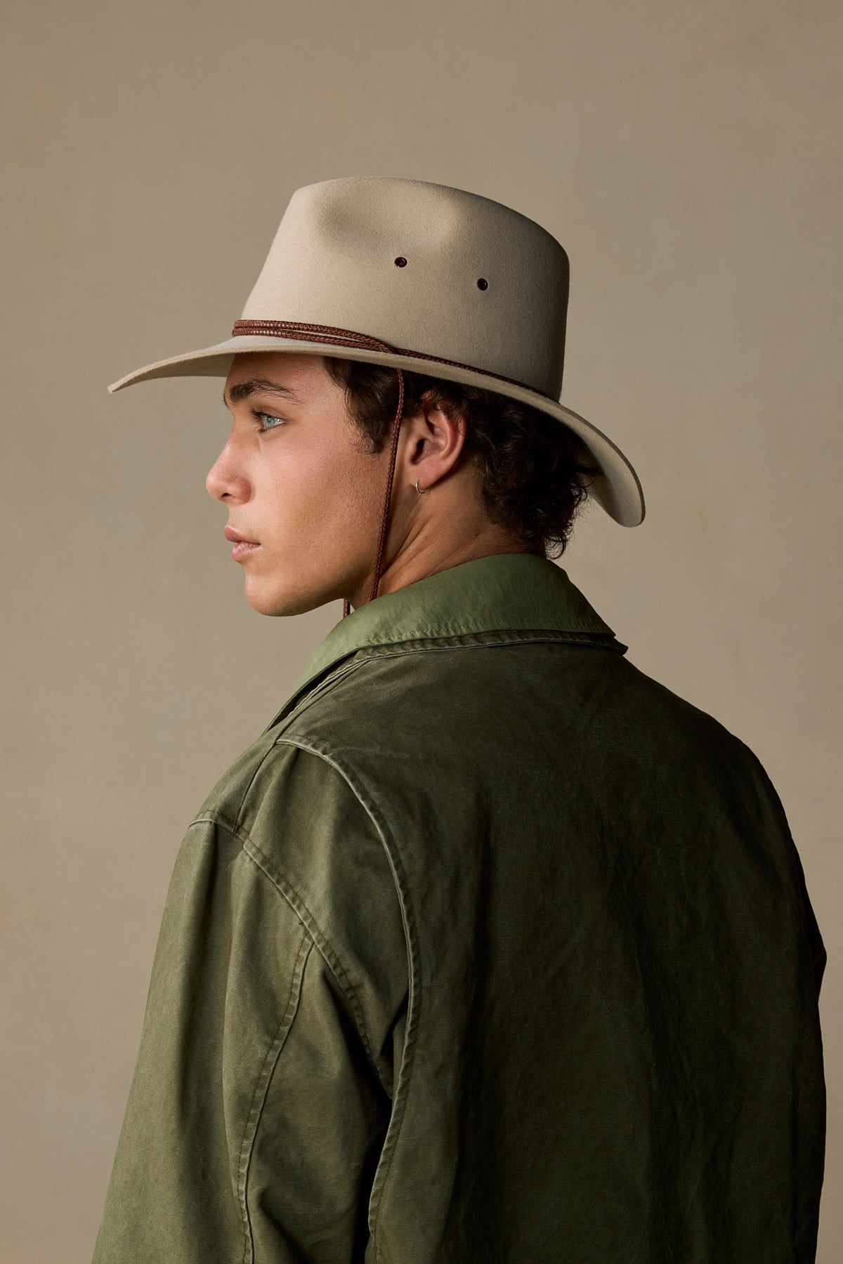 Man wearing sand-colored felt cowboy hat with brown chin strap and olive green jacket, profile view