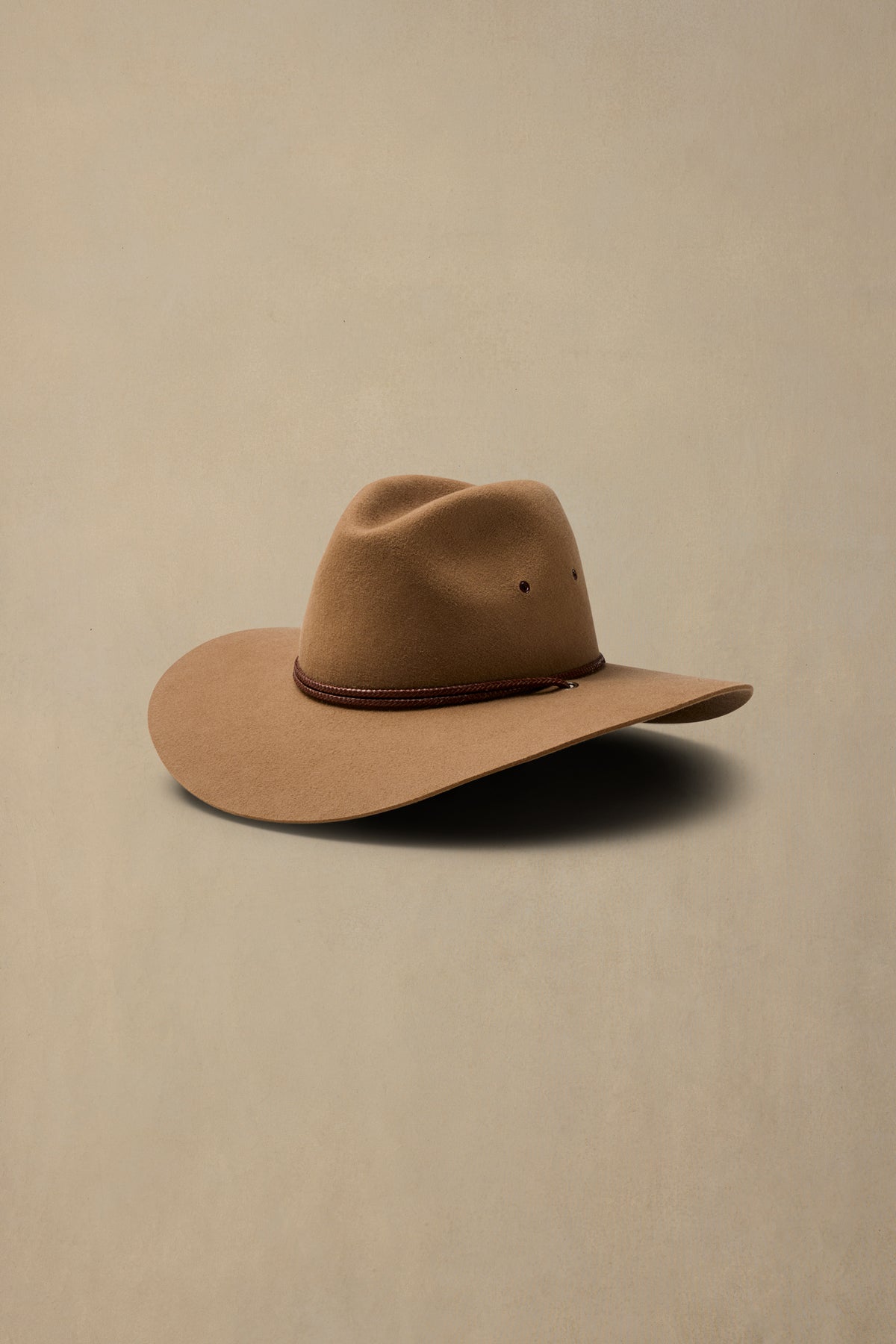 Tan rabbit fur cowboy hat with brown cord band on neutral beige background