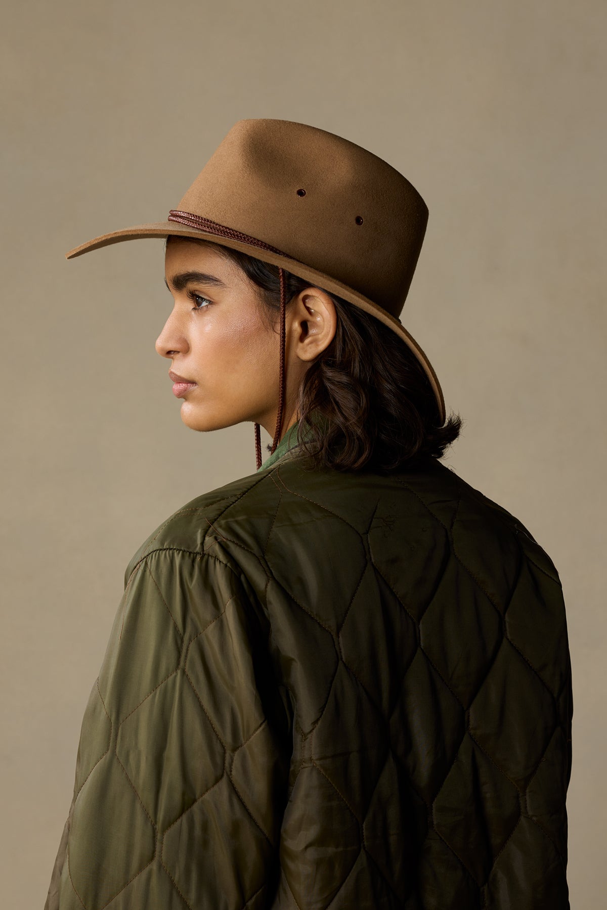 Tan rabbit fur cowboy hat with chin strap worn by woman in olive quilted jacket, side profile view