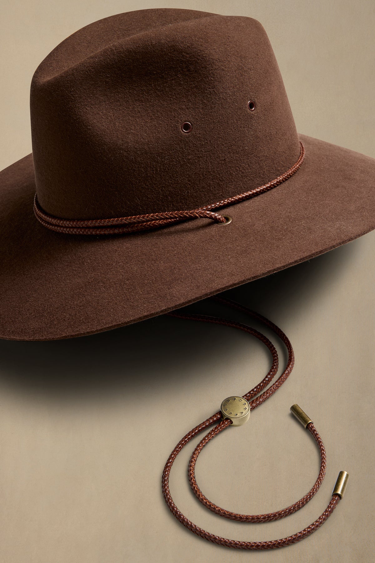 Brown rabbit fur traveller hat with braided leather cord and chin strap detail on neutral background