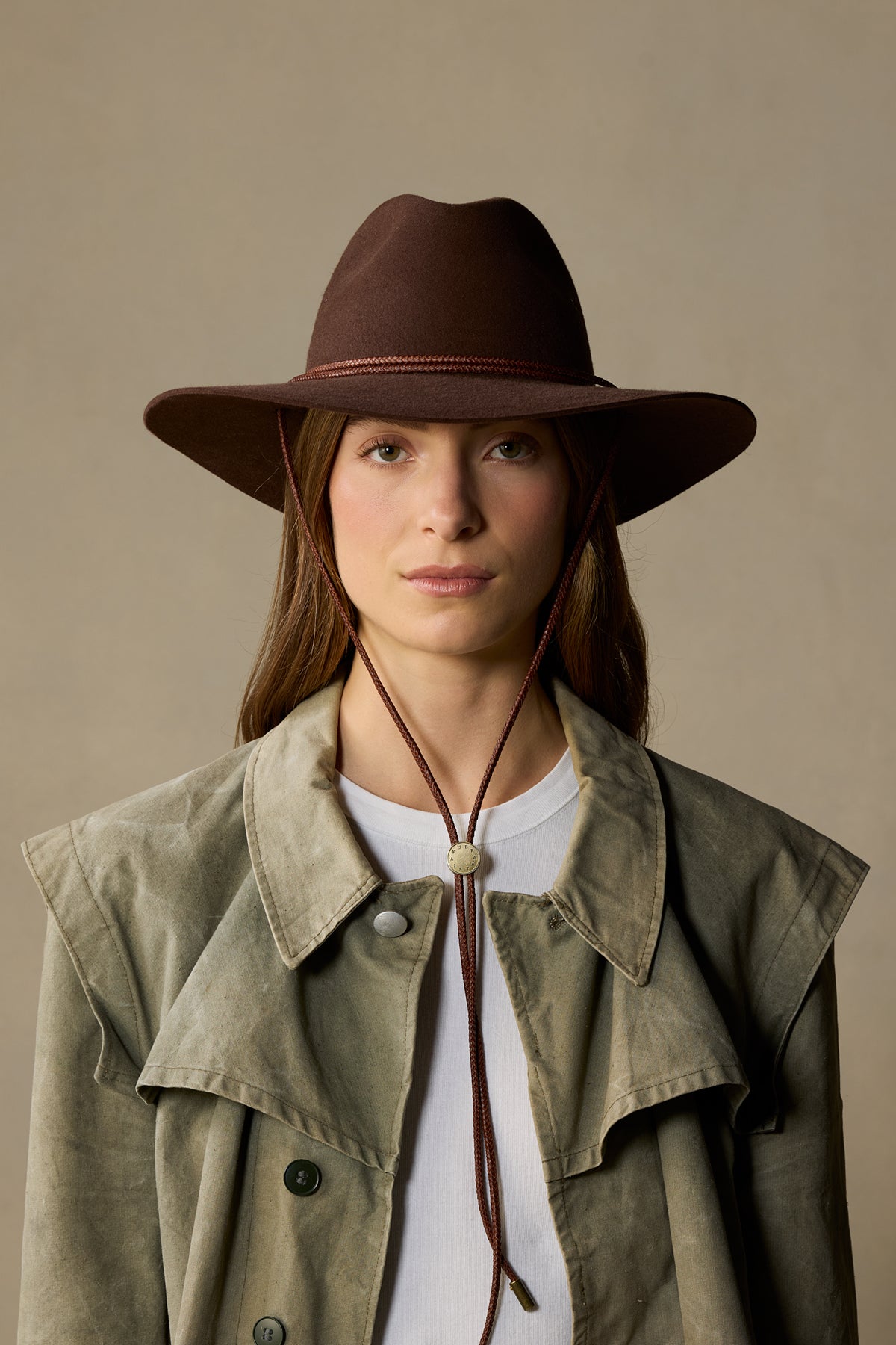 Brown felt cowboy hat with braided band on woman wearing tan jacket and white shirt against neutral background
