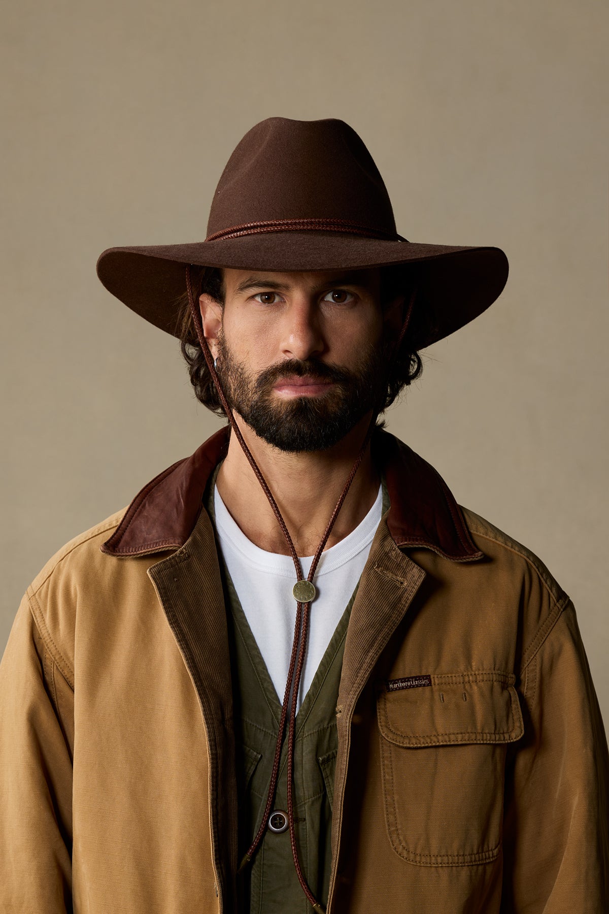 Brown fur felt cowboy hat with leather band and chin strap on bearded man in tan jacket