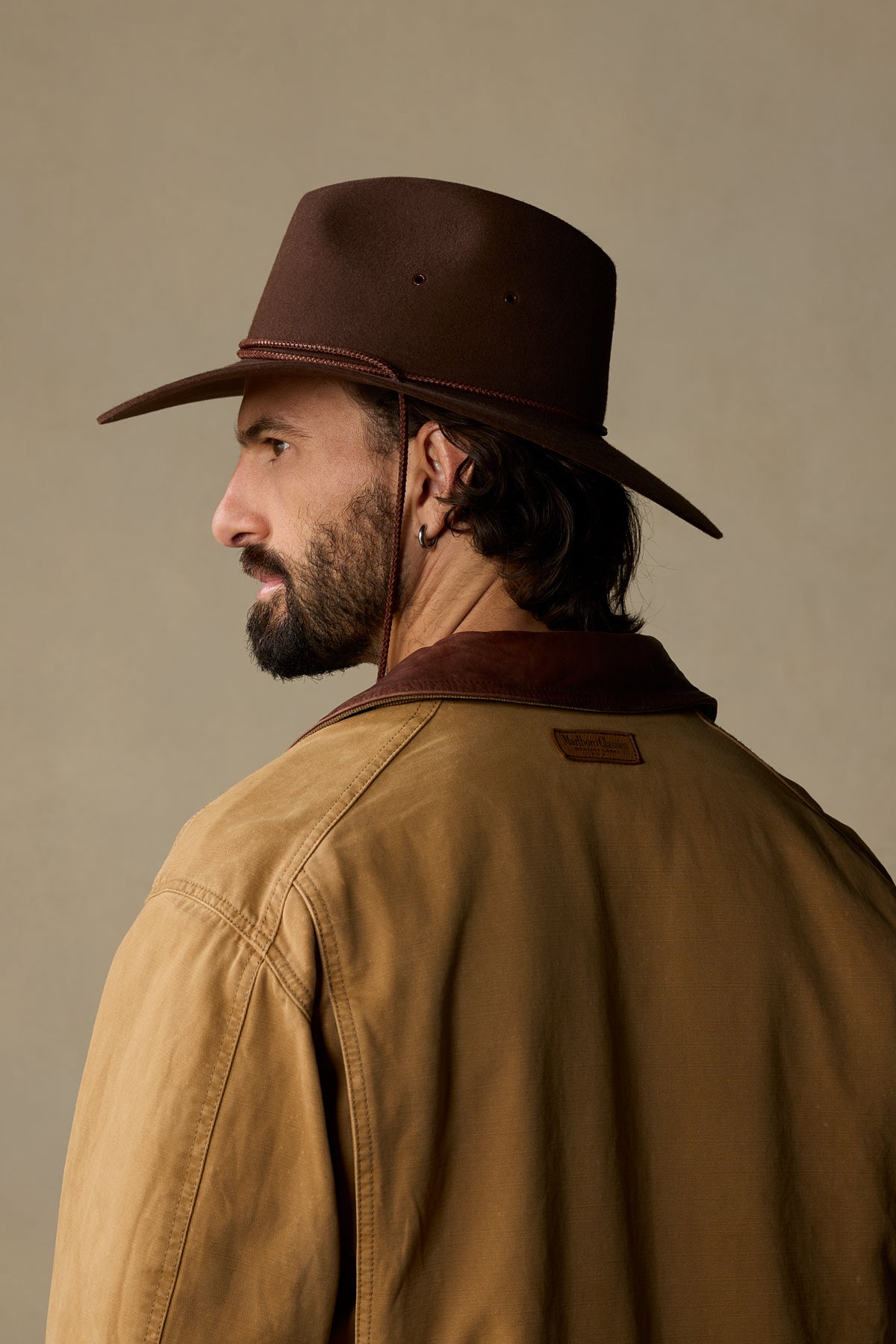 Brown felt traveller hat with chin strap on bearded man wearing tan jacket, side profile view