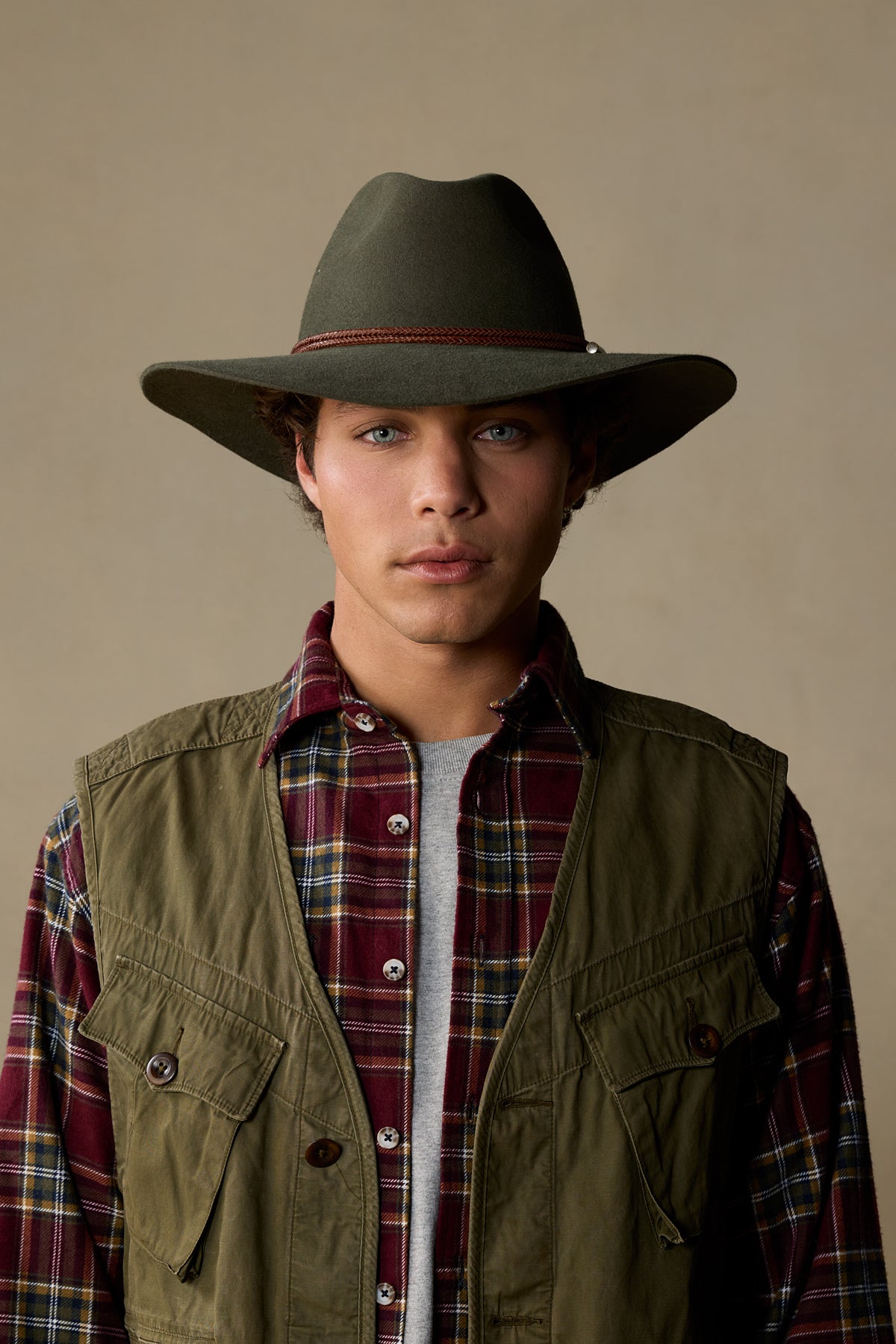 Man wearing olive green vest over burgundy plaid shirt and gray cowboy hat against neutral background