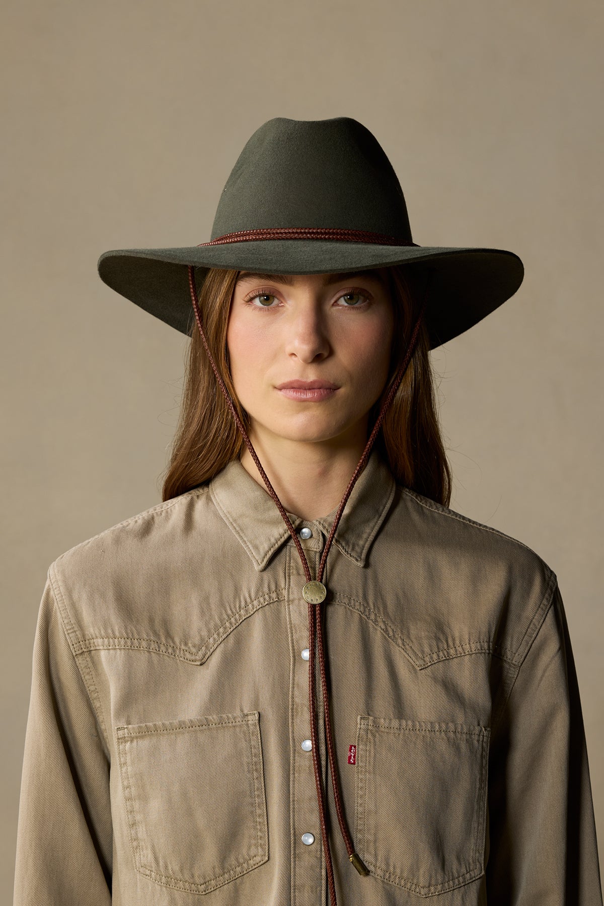 Woman wearing fern green fur felt cowboy hat with burgundy chin cord and tan denim shirt against neutral background