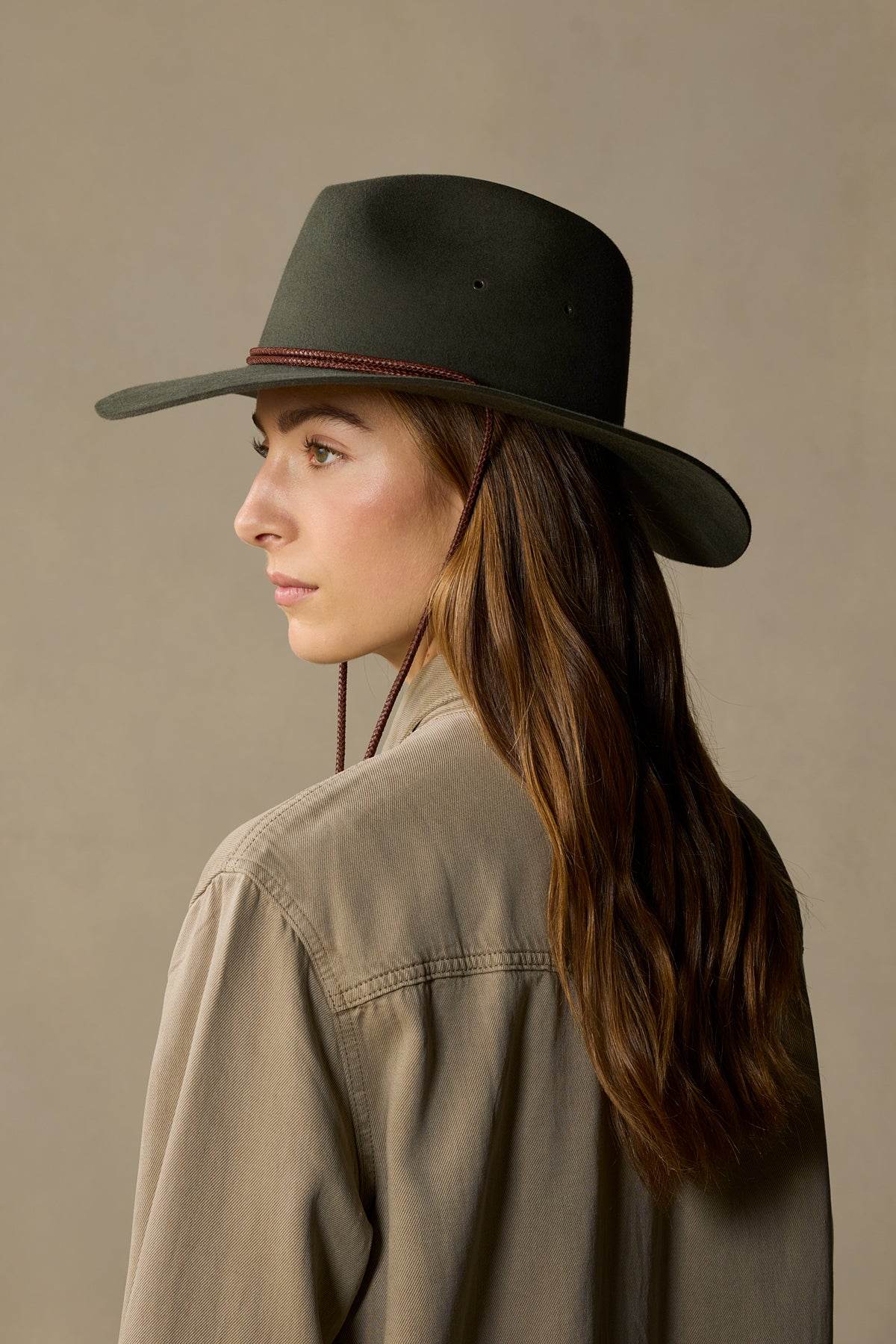Woman wearing olive green rabbit fur traveller hat with chin strap, turned profile view against neutral background