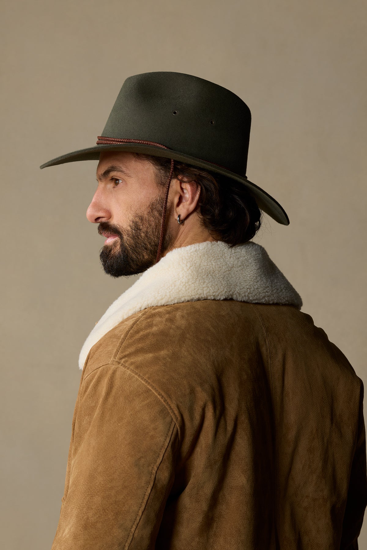 Man wearing olive green cowboy hat with chin strap and tan suede jacket with sherpa lining, side profile view