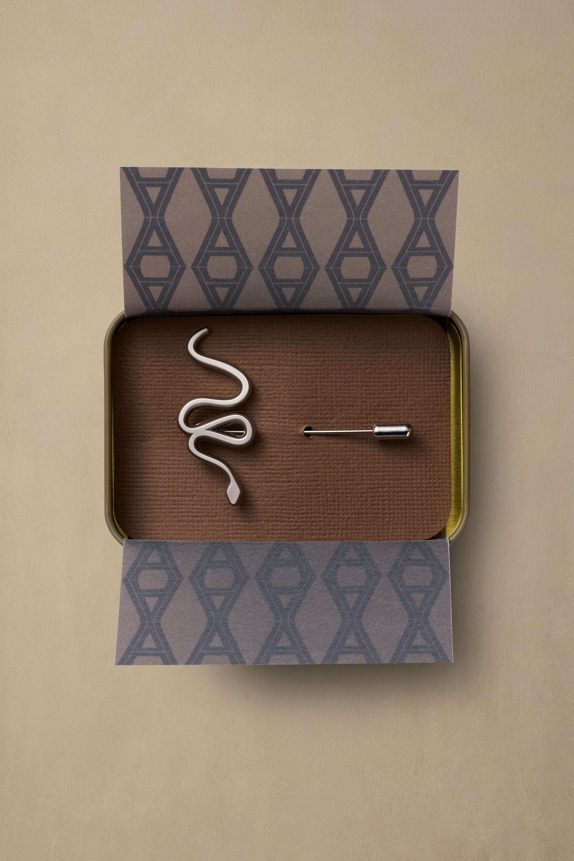 Red Belly Snake pewter pin in coiled design, displayed in open bronze tin with patterned paper packaging on neutral background