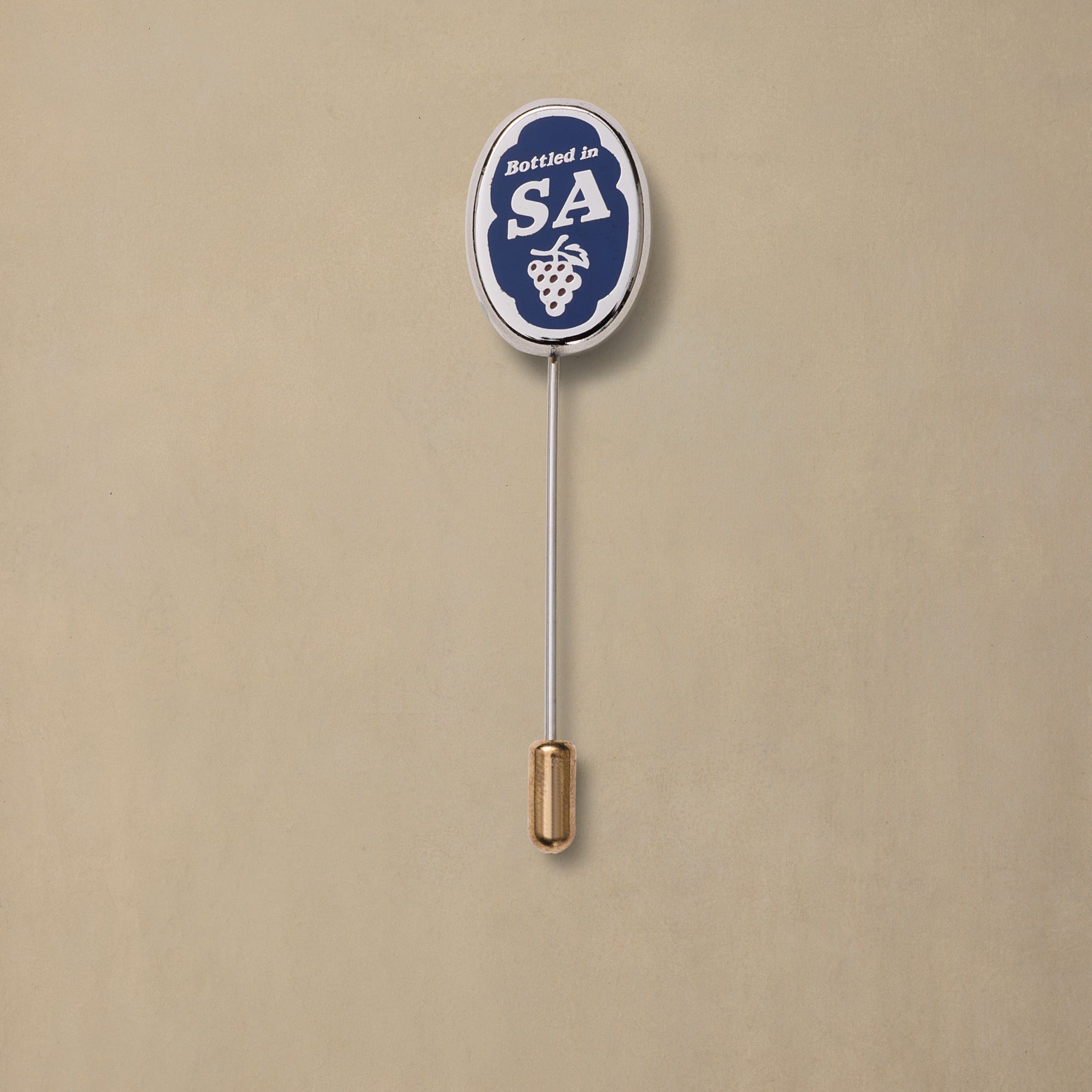 Navy and white enamel pin badge with "Bottled in SA" text and grape bunch design, gold-tone backing, on beige background