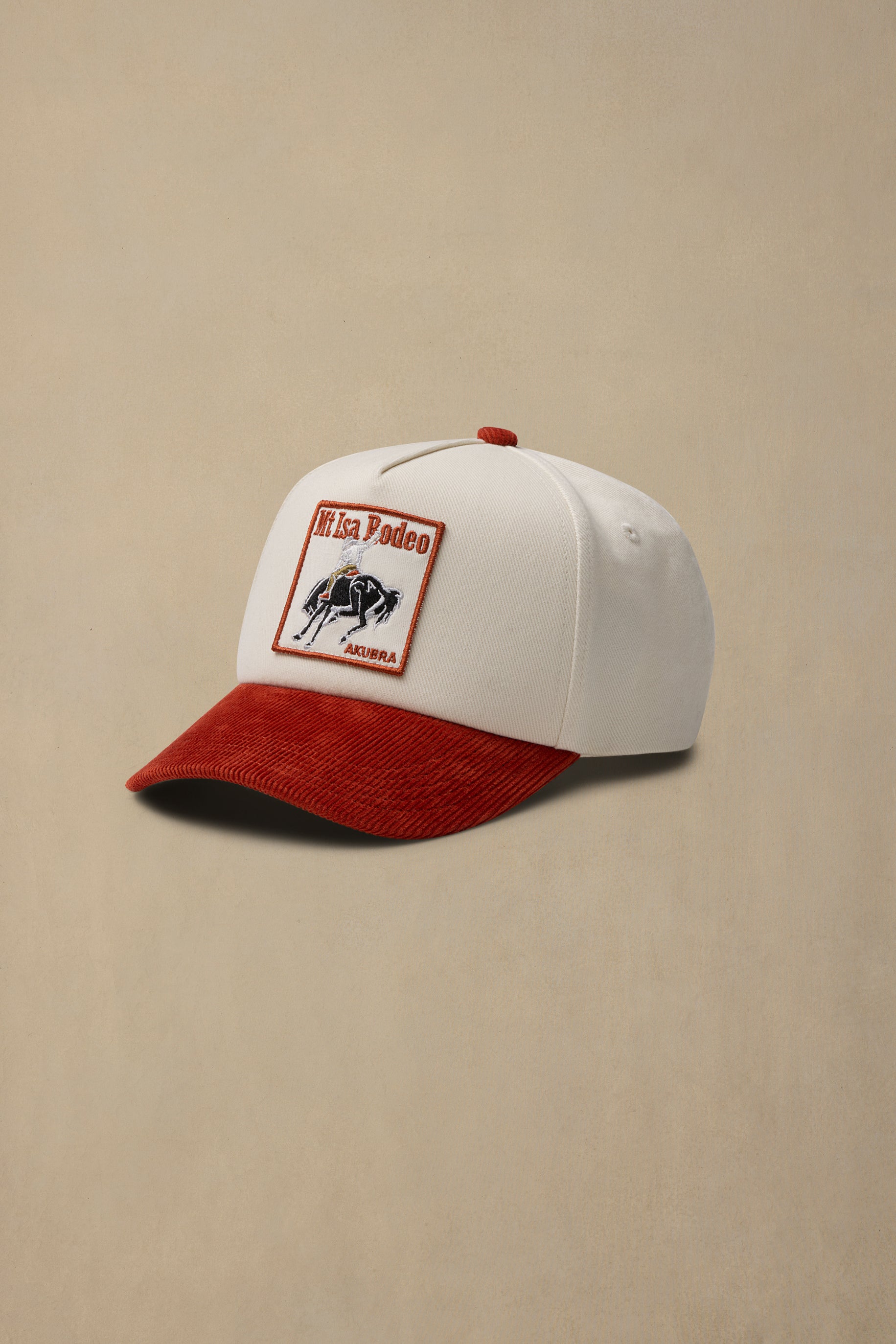 A white baseball cap with a red corduroy brim and a rectangular 'Mt Isa Rodeo 2026' patch on the front, set against a solid beige background.