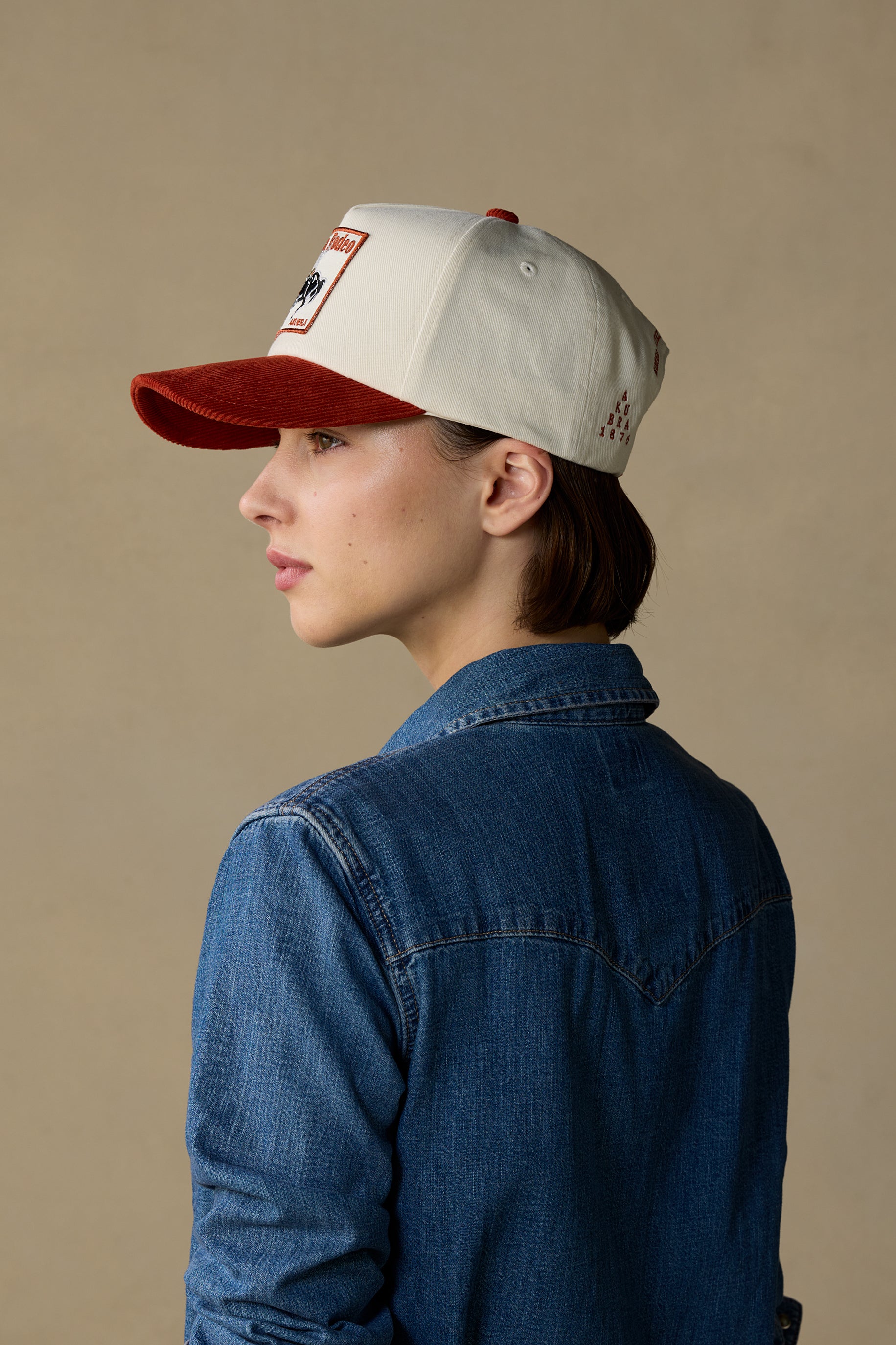 A woman in profile wearing a light gray and red baseball cap with a denim shirt, set against a plain beige background.