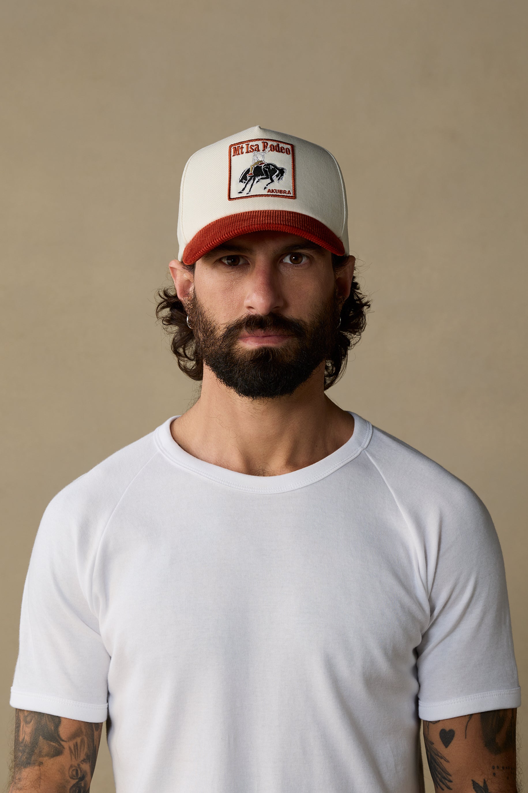 A man with a beard and tattoos wearing a white crew-neck t-shirt and a white trucker hat with a red brim and a 'Mt Isa Rodeo' logo patch, posed against a solid beige background.