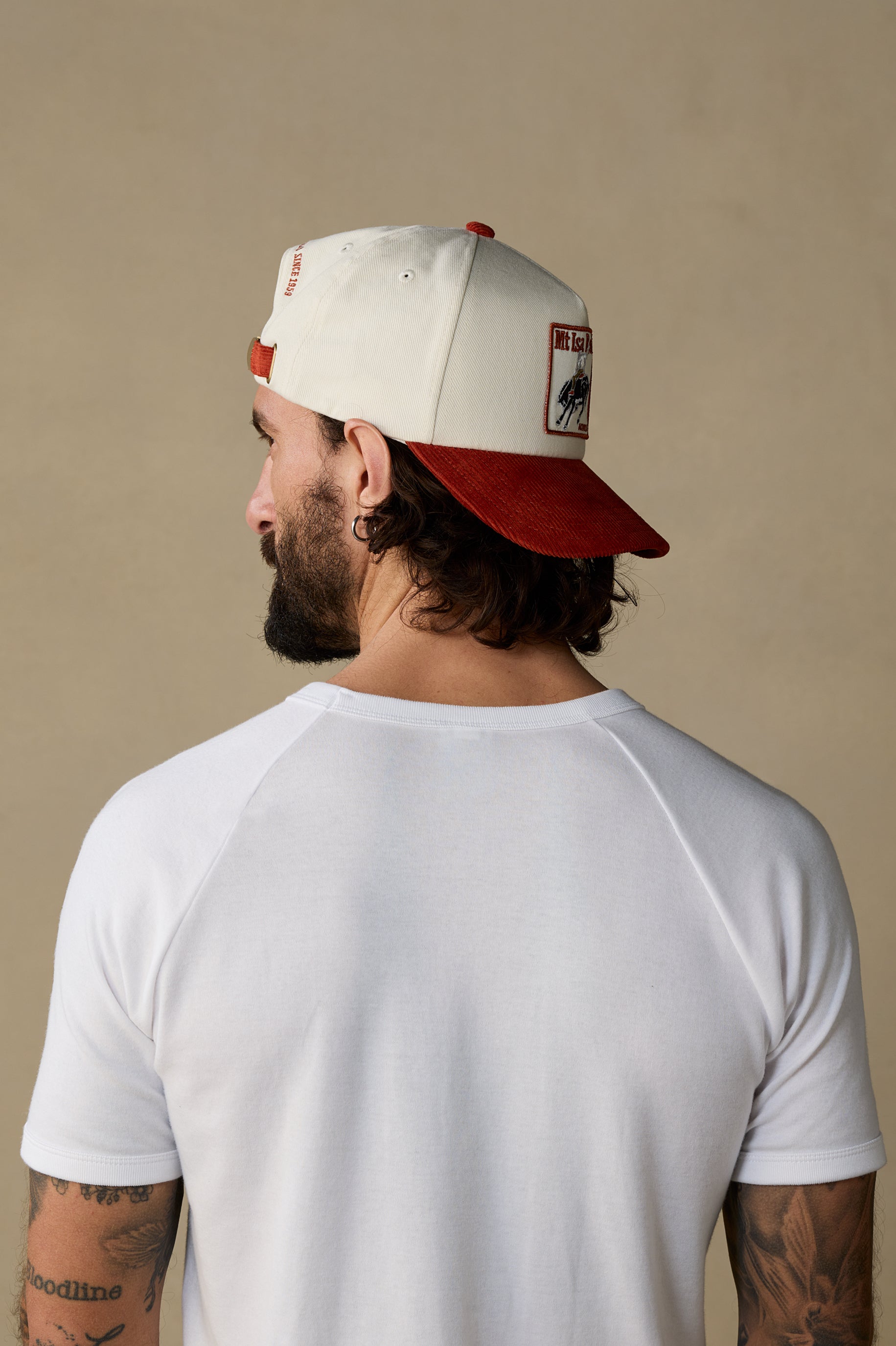 A man shown from the back wearing a white and red baseball cap and a white raglan t-shirt against a solid beige background.