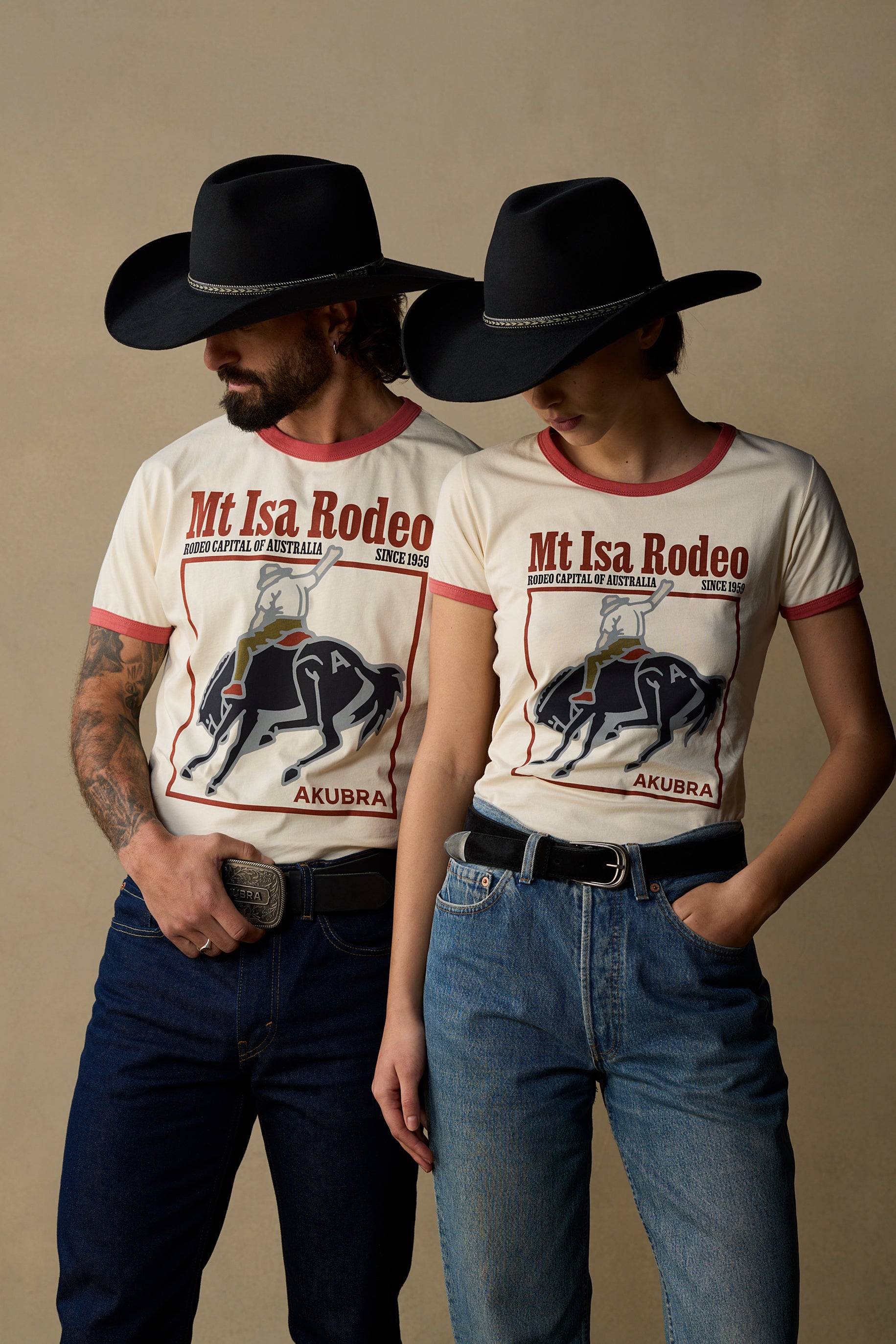 Two models wearing matching off-white ringer t-shirts with red trim and a graphic print of 'Mt Isa Rodeo', styled with black cowboy hats, dark blue jeans, and black belts against a neutral beige background.