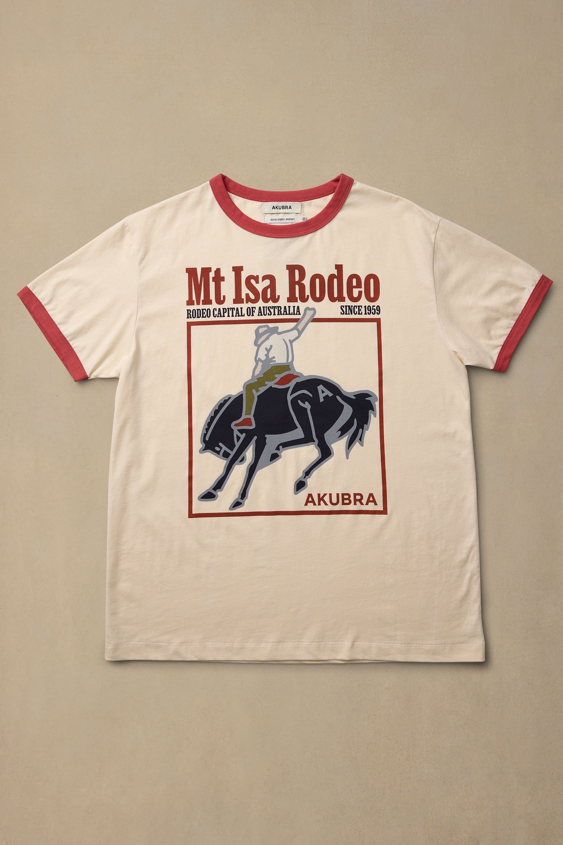 An off-white ringer t-shirt with red trim on the collar and sleeves, featuring a graphic print of a cowboy riding a black horse under the text 'Mt Isa Rodeo' and 'AKUBRA'.