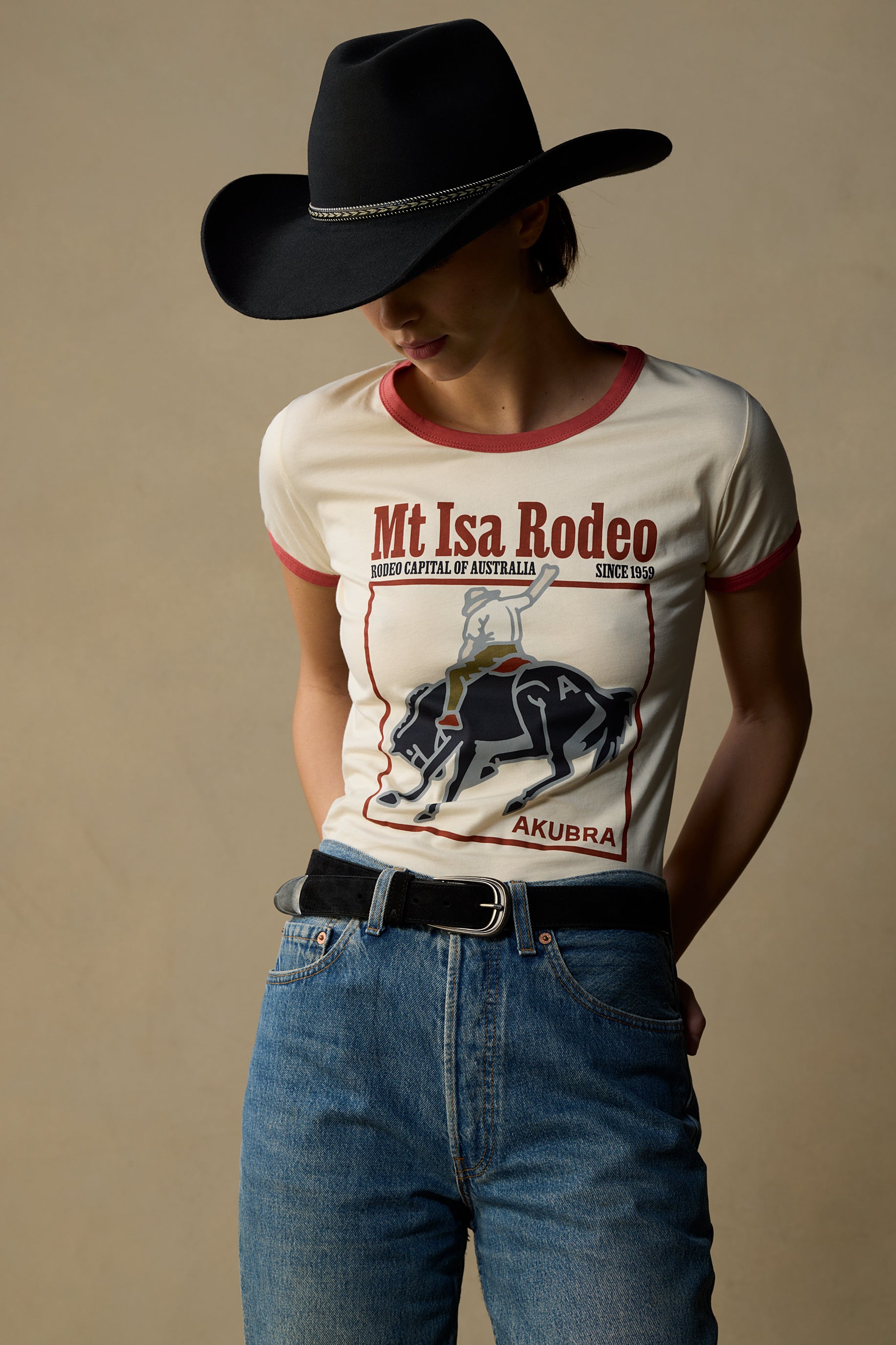 A model wearing a cream-colored ringer t-shirt with red trim and a 'Mt Isa Rodeo' graphic, styled with blue jeans, a black belt, and a black cowboy hat against a solid beige background.