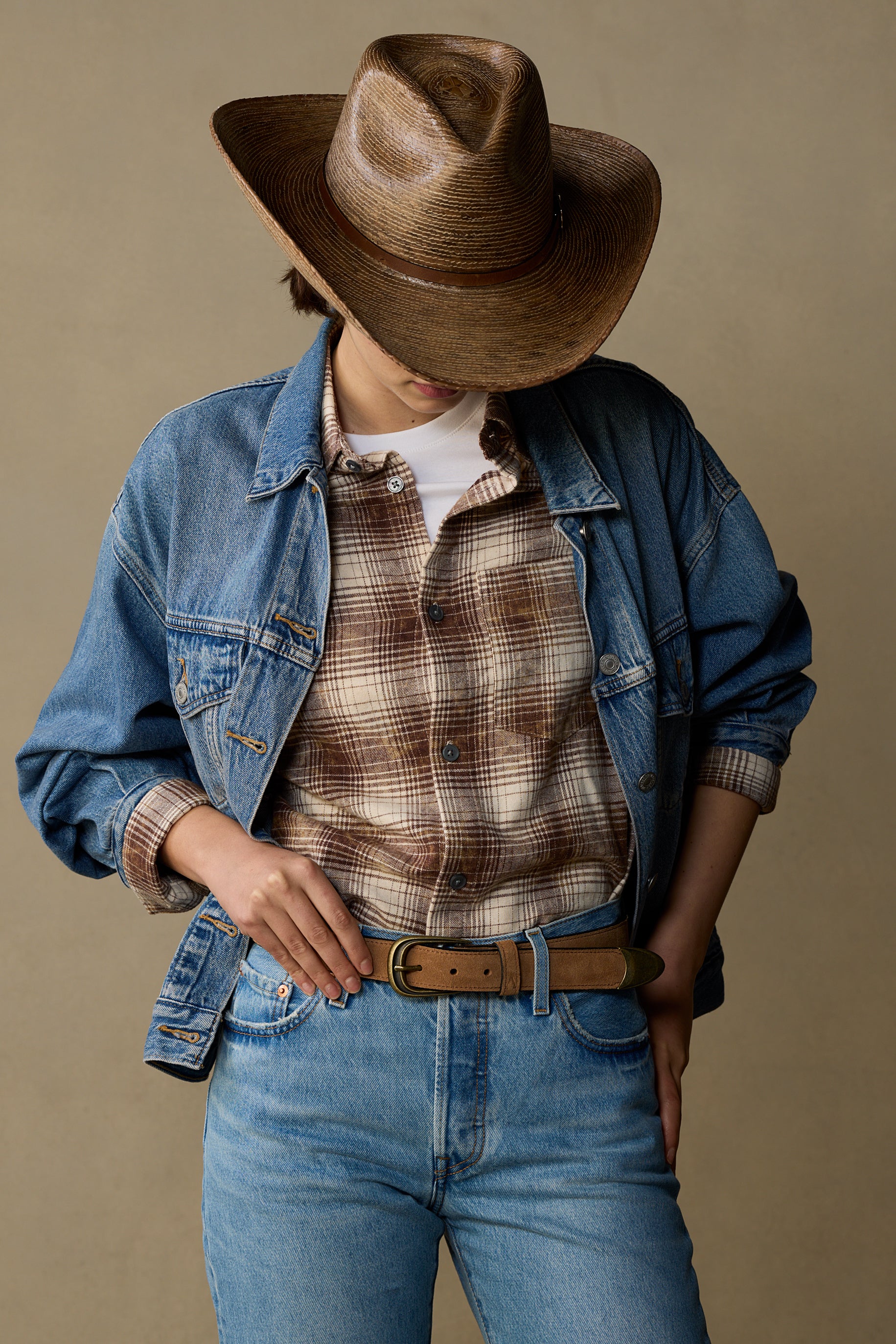 Tan suede leather belt with brass buckle worn with denim jacket and plaid shirt on neutral background