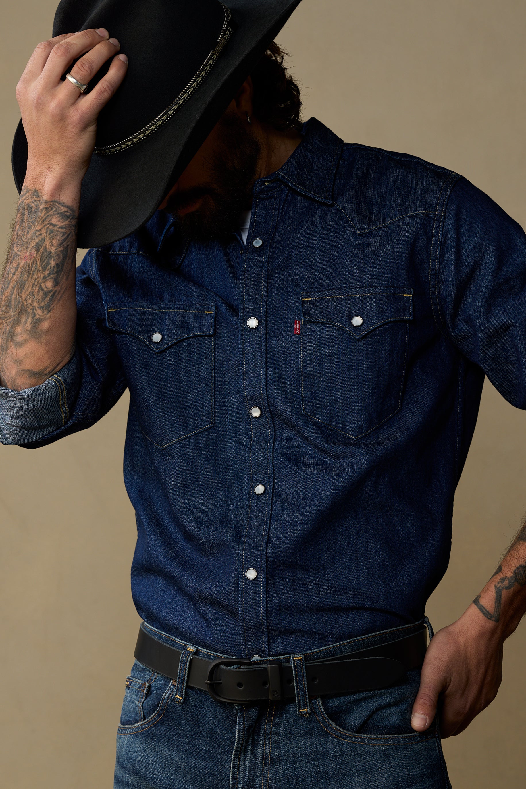 Black leather belt worn with dark denim shirt and cowboy hat against neutral background
