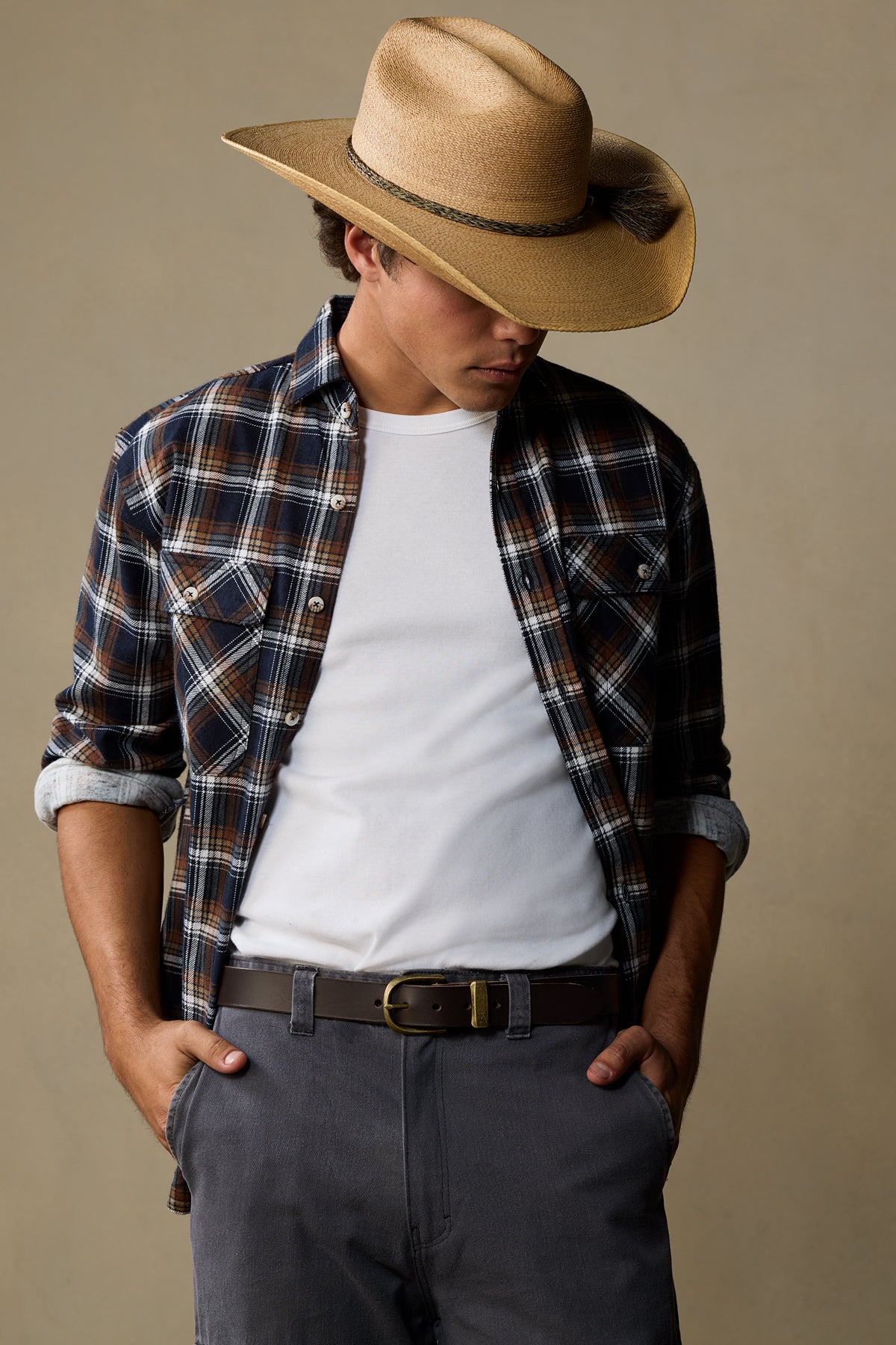 Tan leather belt with brass buckle worn with plaid shirt and cowboy hat on neutral background