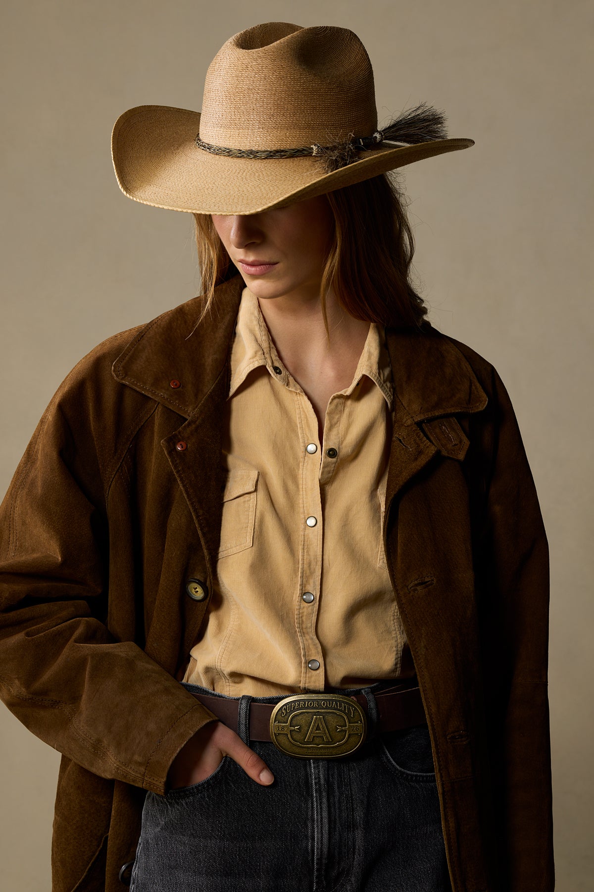 Chocolate brown leather belt with brass badge buckle worn with tan shirt and brown jacket on neutral background
