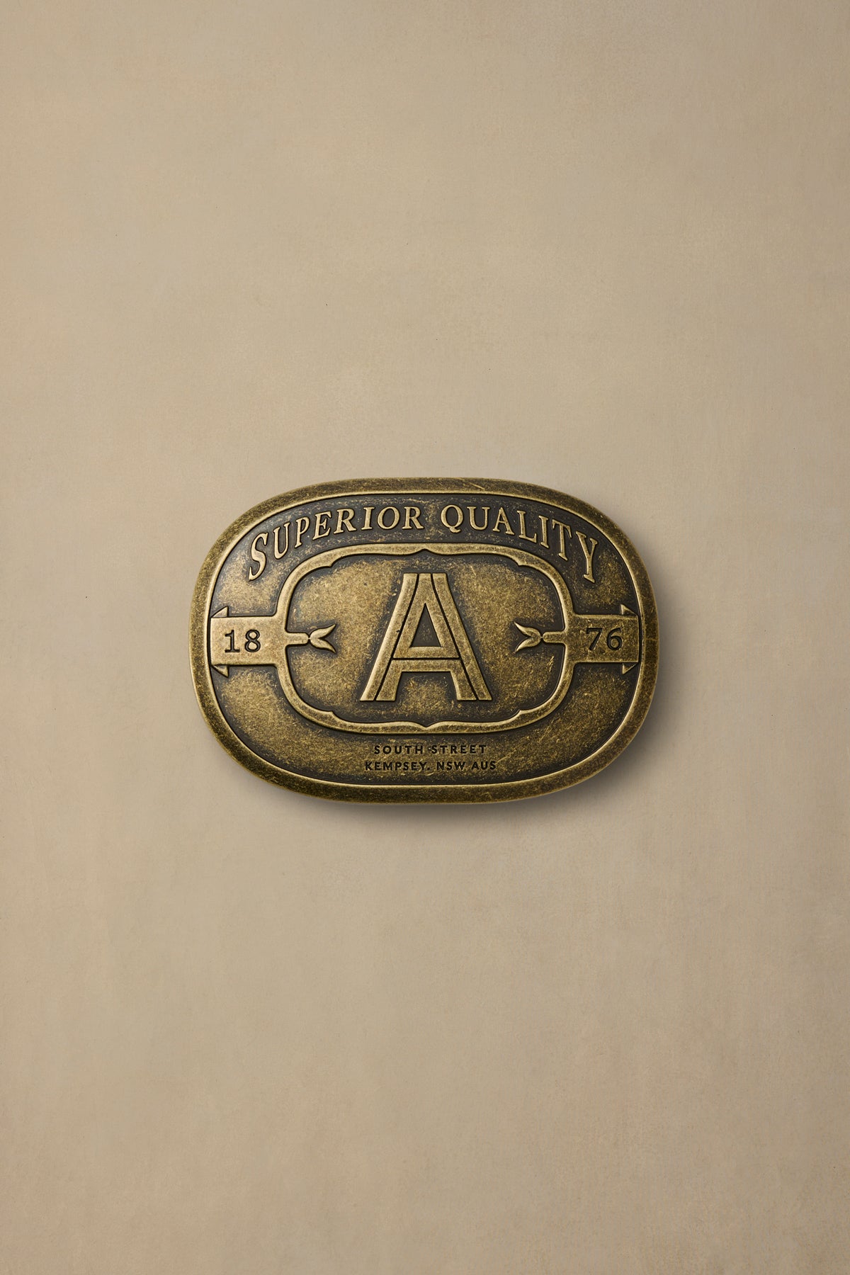 Vintage brass belt buckle with "A" initial and "Superior Quality" text from 1876, displayed against neutral beige background