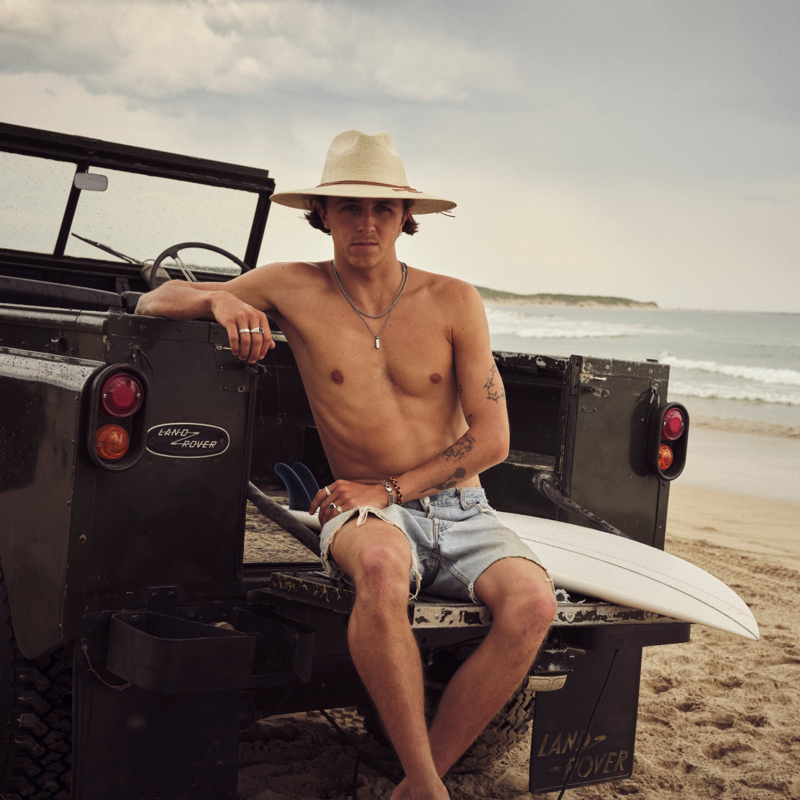 Man sitting on a Land Rover on a beach with a surfboard wearing an Akubra straw hat
